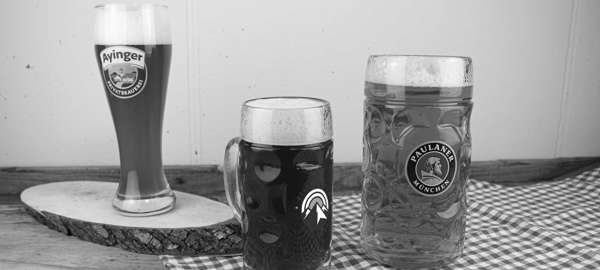 Verschiedene personalisierte Biergläser und Glaskrüge mit individueller Gravur auf Holzbrett und karierter Tischdecke.