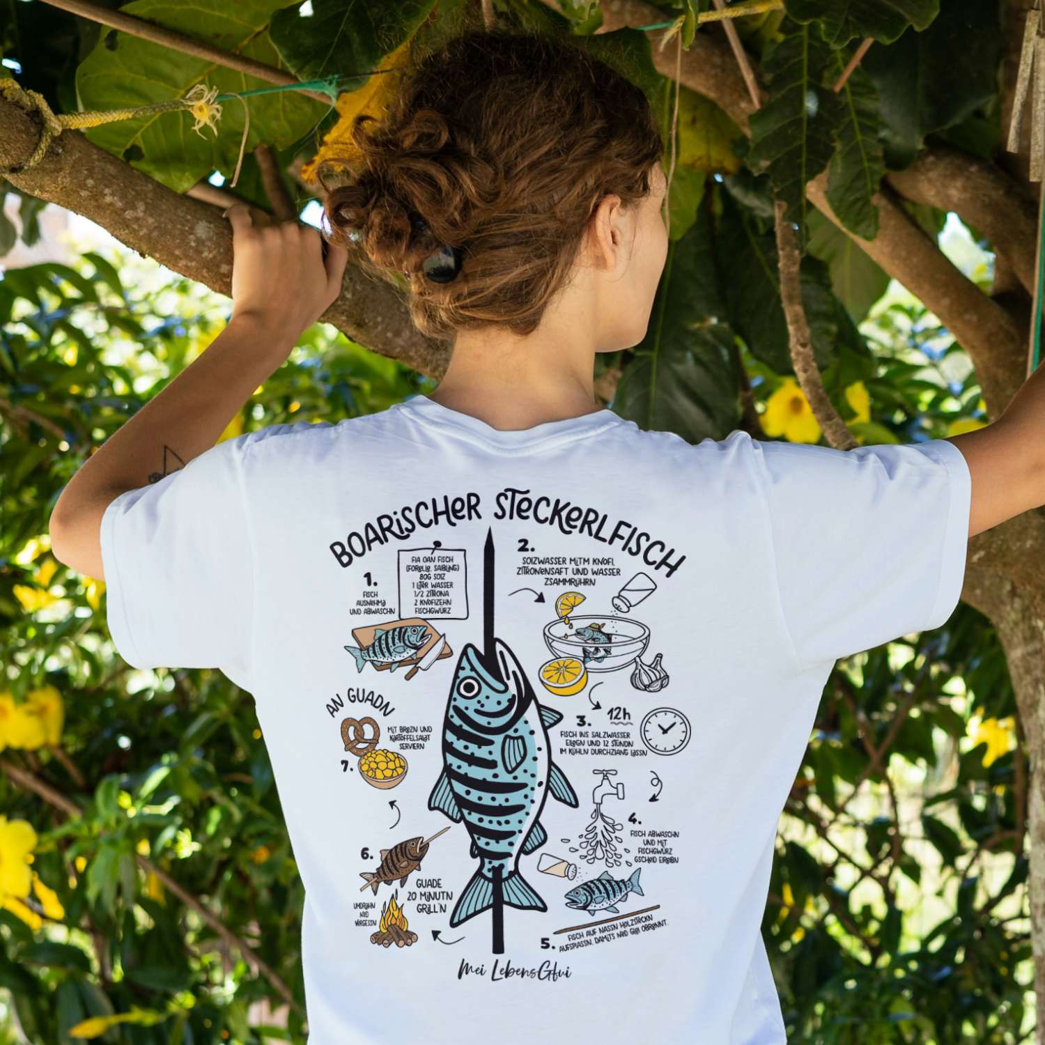 Rückenansicht eines Damen T-Shirts "Boarischer Steckerlfisch" mit Rezeptanleitung unter Bäumen.