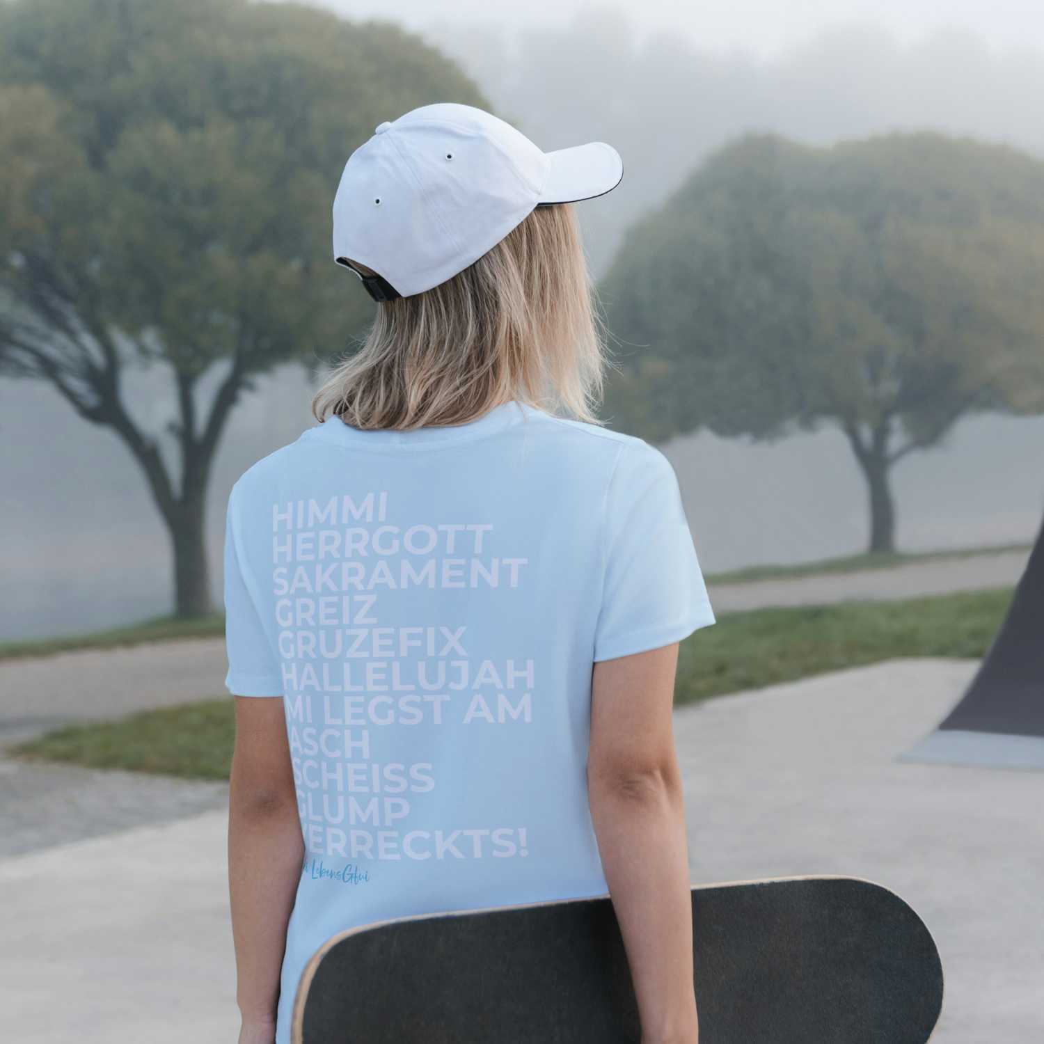 Rückenansicht einer Frau mit weißem Damen T-Shirt Himmi Herrgott, Skateboard haltend, leichte Hügellandschaft.