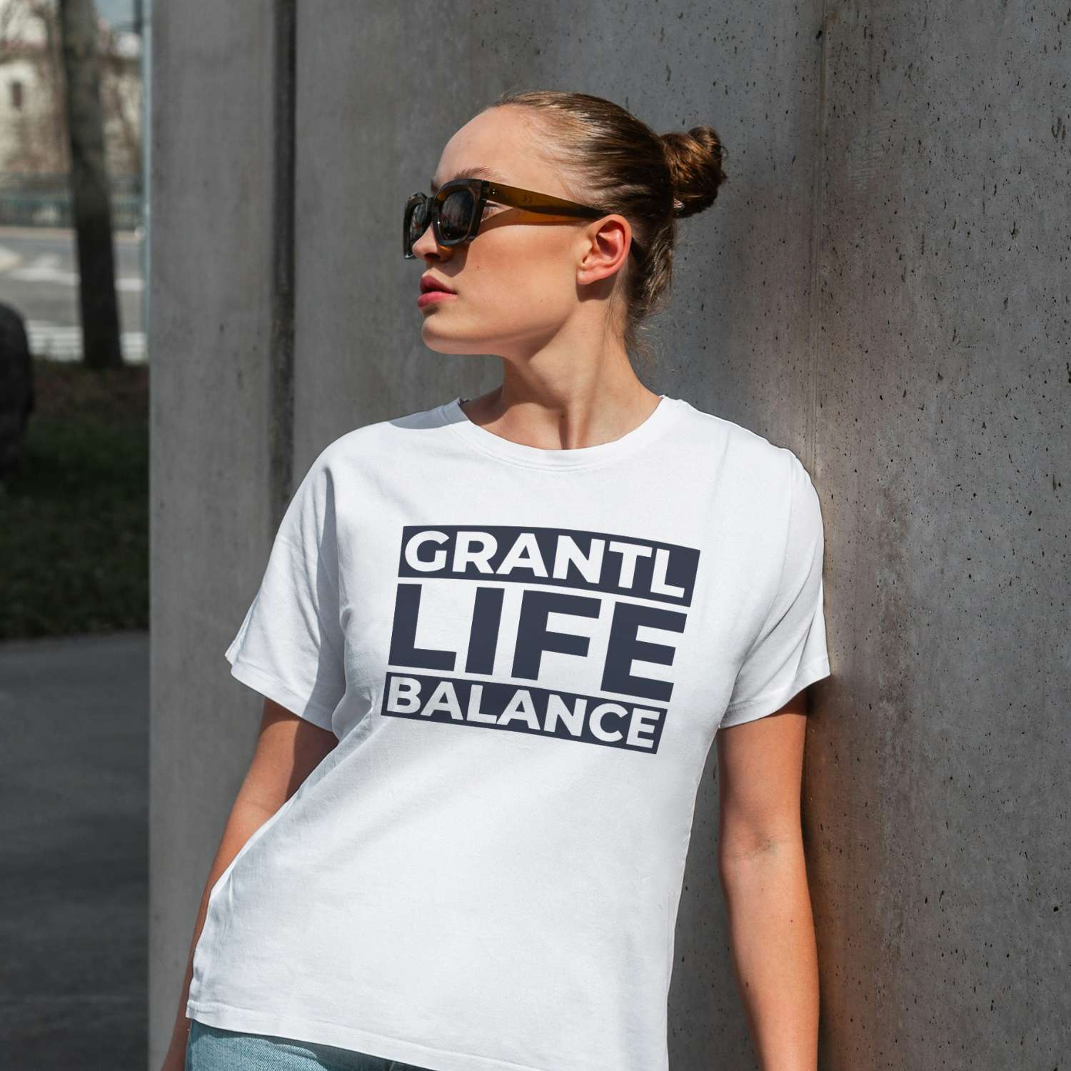 Junge Frau trägt weißes Damen T-Shirt Grantl Life Balance mit schwarzem Druck, Sonnenbrille und Dutt, lehnt lässig an Wand.