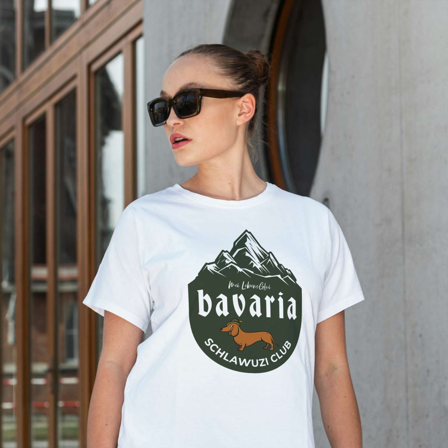 Model trägt weißes Damen T-Shirt Bavaria Schlawuzi Club mit Dackel-Motiv und Alpenpanorama, lässiger Look.