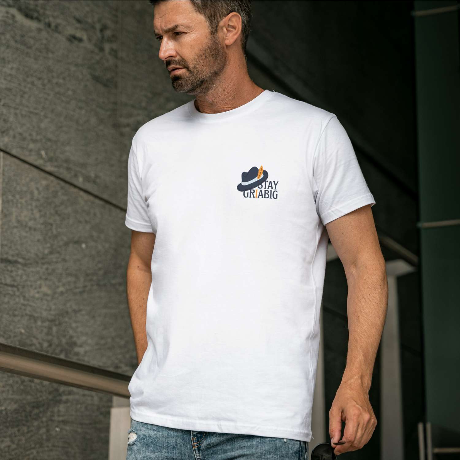 Mann trägt weißes Herren T-Shirt Stay Griabig mit dunkelblauem Hut-Logo und dezenten Nietenjeans.
