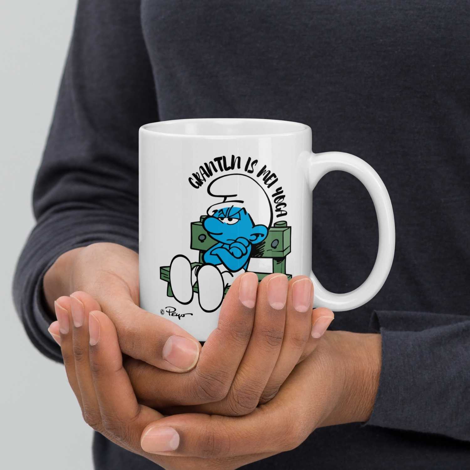 Weiße ®Die Schlümpfe Tasse "Grantln is mei Yoga" mit mürrischem Schlumpf in den Händen einer Person.