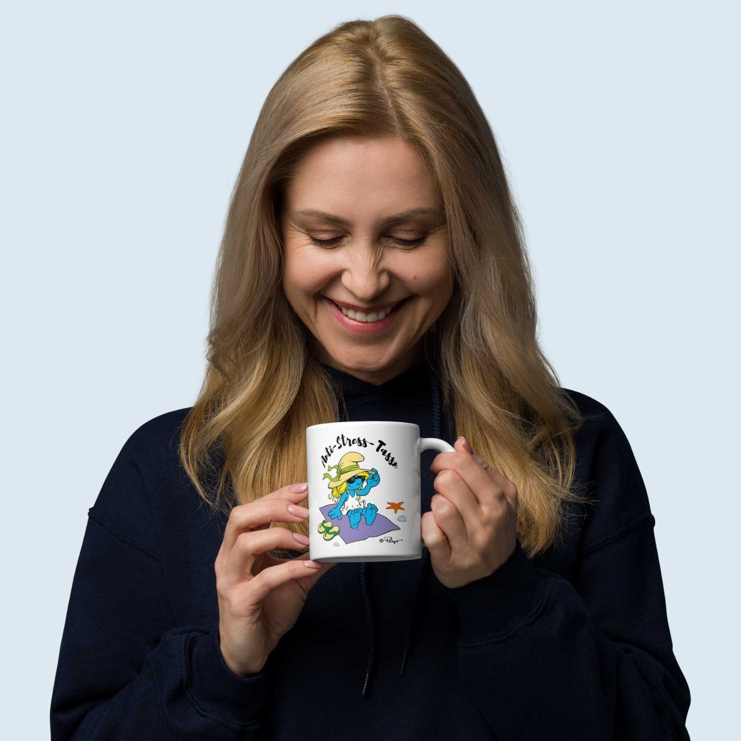 Lächelnde Frau hält eine weiße ®Die Schlümpfe Tasse Anti-Stress Damen mit einem coolen Schlumpf auf einem Handtuch.