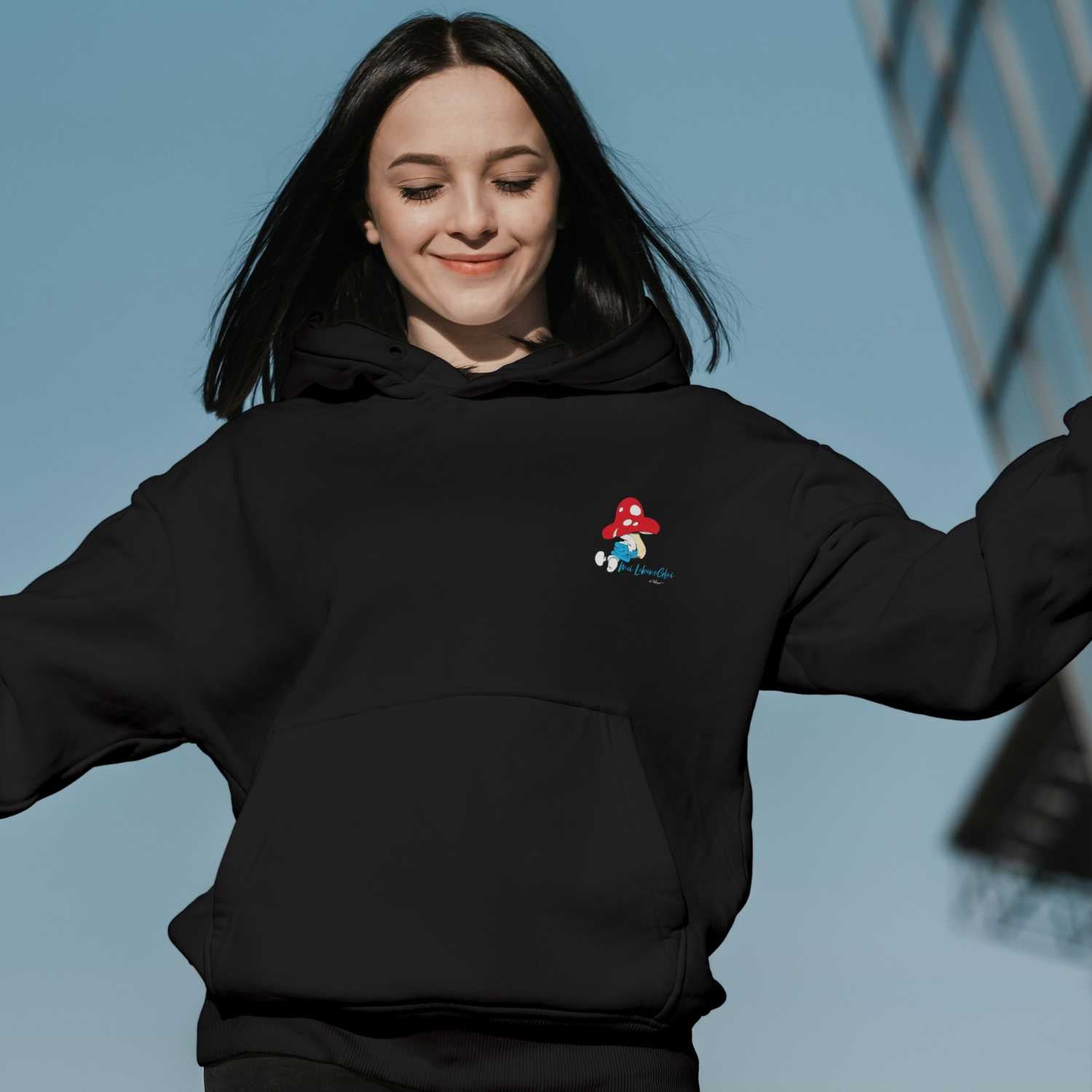 Lächelnde Frau trägt schwarzen Damen Hoodie mit "Die Schlümpfe" Motiv und Pilz-Stickerei, blauem Himmel.