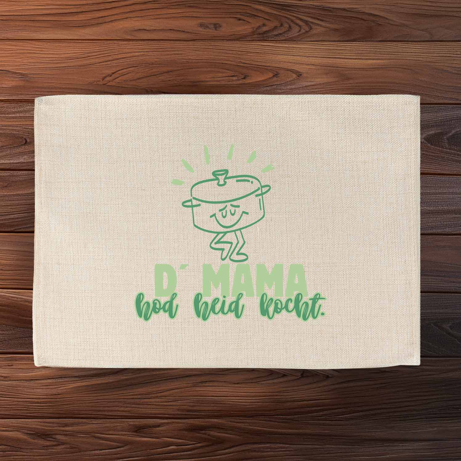 Helles Leinen-Tischset D´Mama hod heid kocht mit grüner Kochtopf-Illustration und Schriftzug auf Holztisch.