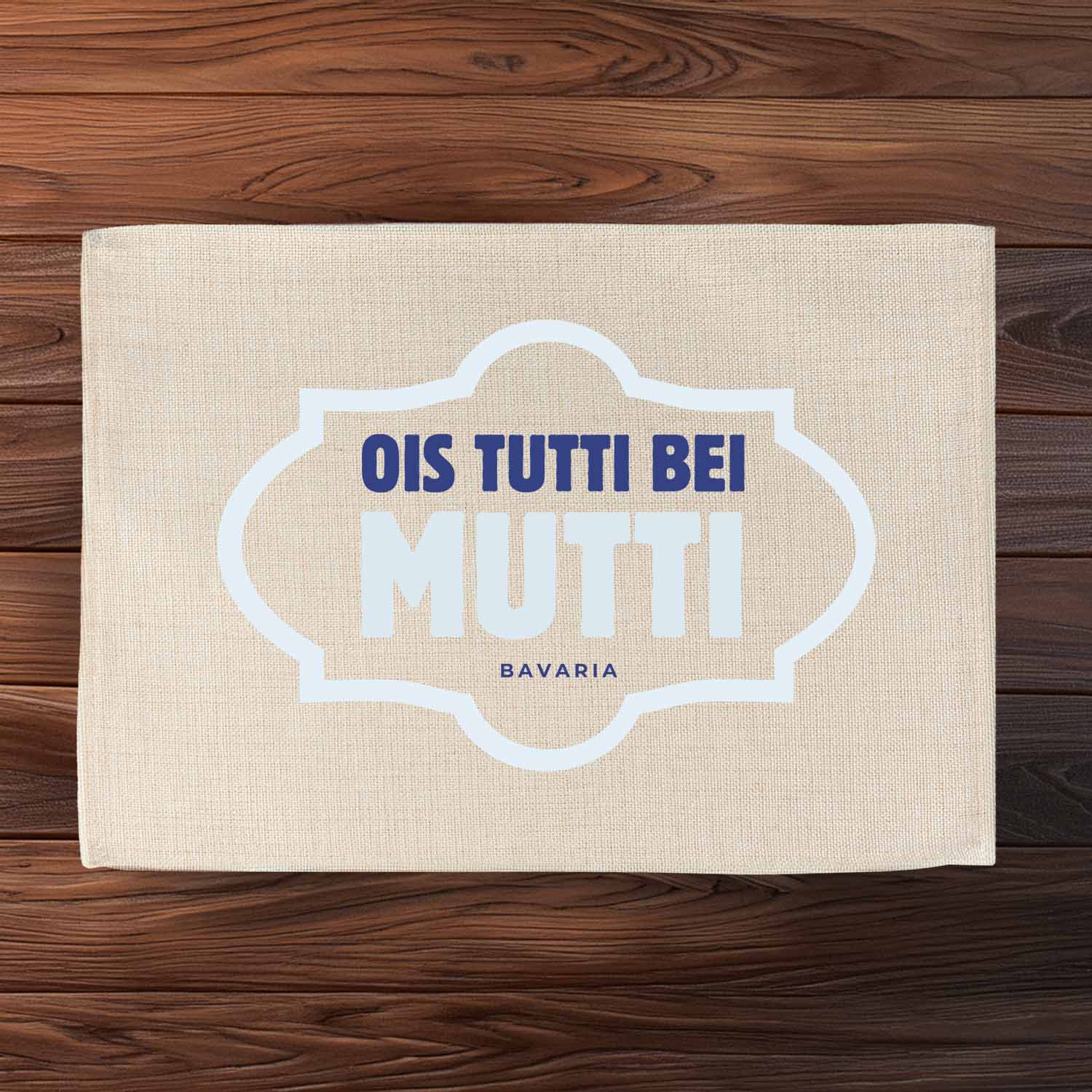 Beiges Tischset Ois Tutti bei Mutti mit blauem bayrischem Schriftzug auf dunklem Holztisch.