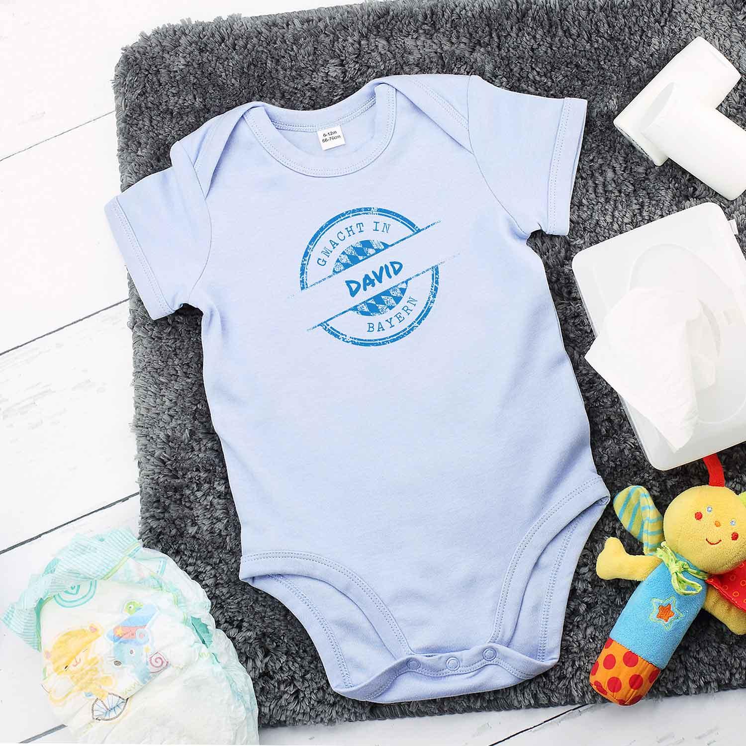 Baby Body "gmacht in Bayern" blau mit individuellem Wunschnamen "David" auf grauer Decke.