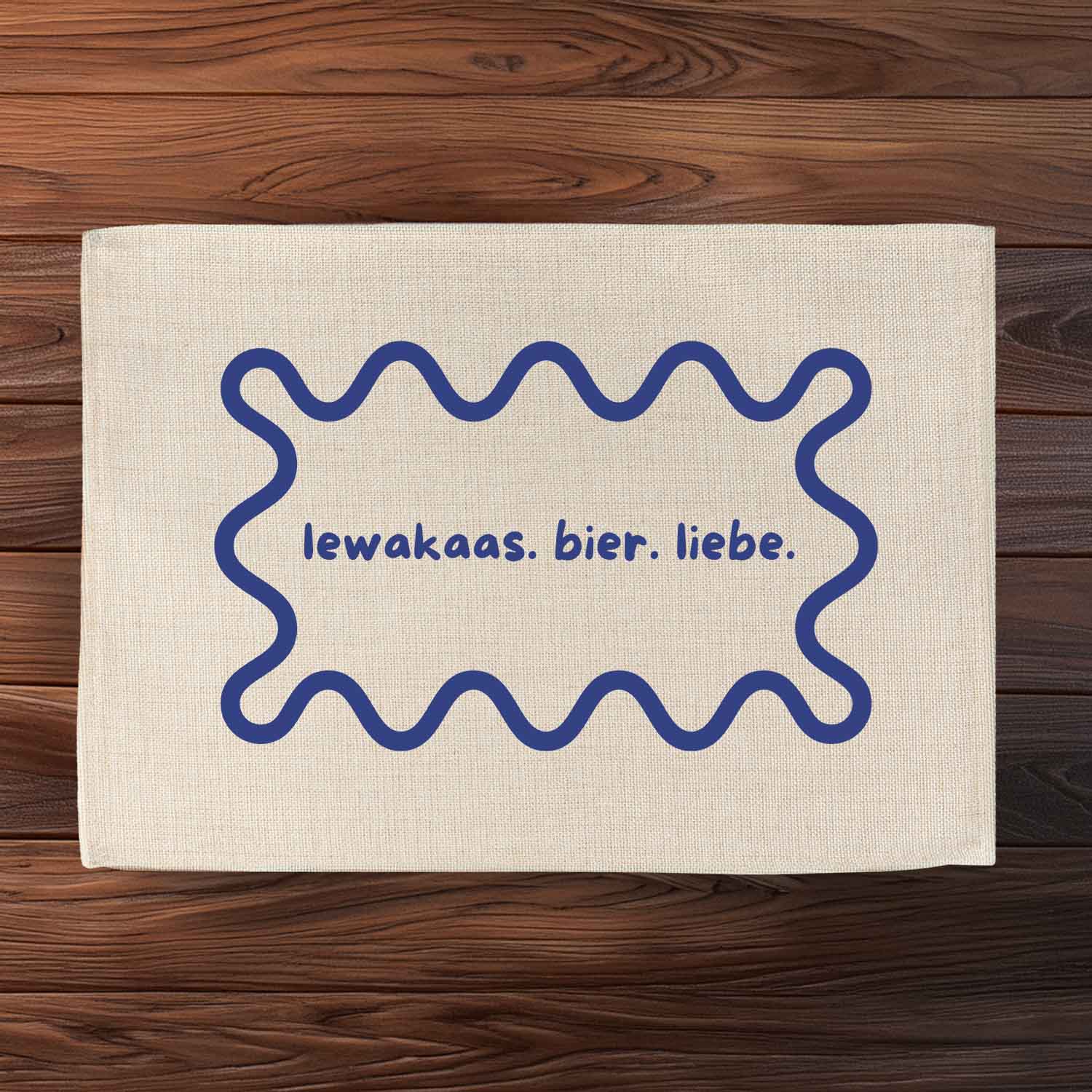 Tischset "lewakaas. bier. liebe." aus hellem Stoff mit blauem Rahmen und Schriftzug auf dunklem Holztisch.