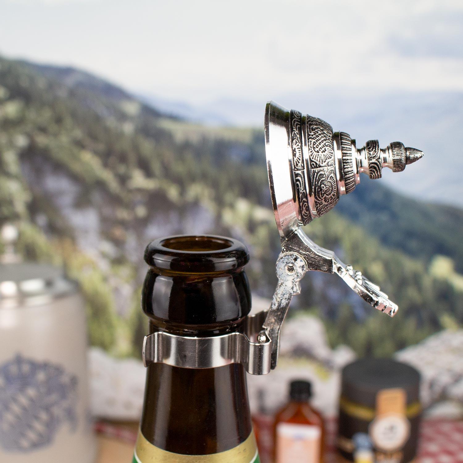 Metallischer Flaschenaufsatz mit Klappdeckel auf einer Bierflasche, traditionell verziert, vor Alpenkulisse.