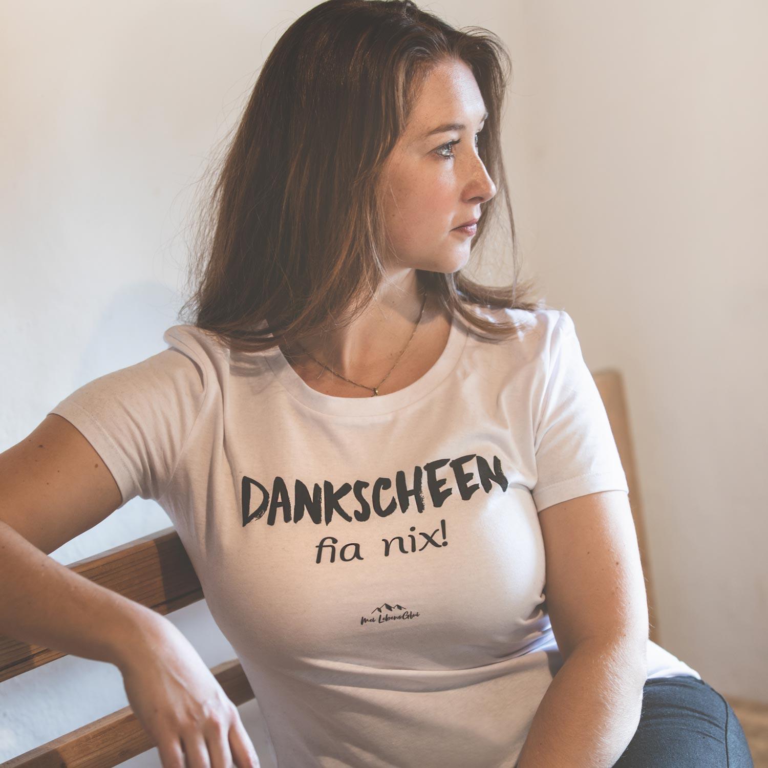 Frau trägt schlichtes, weißes Damen T-Shirt "Dankscheen - fia nix!" lässig sitzend auf Holzbank.