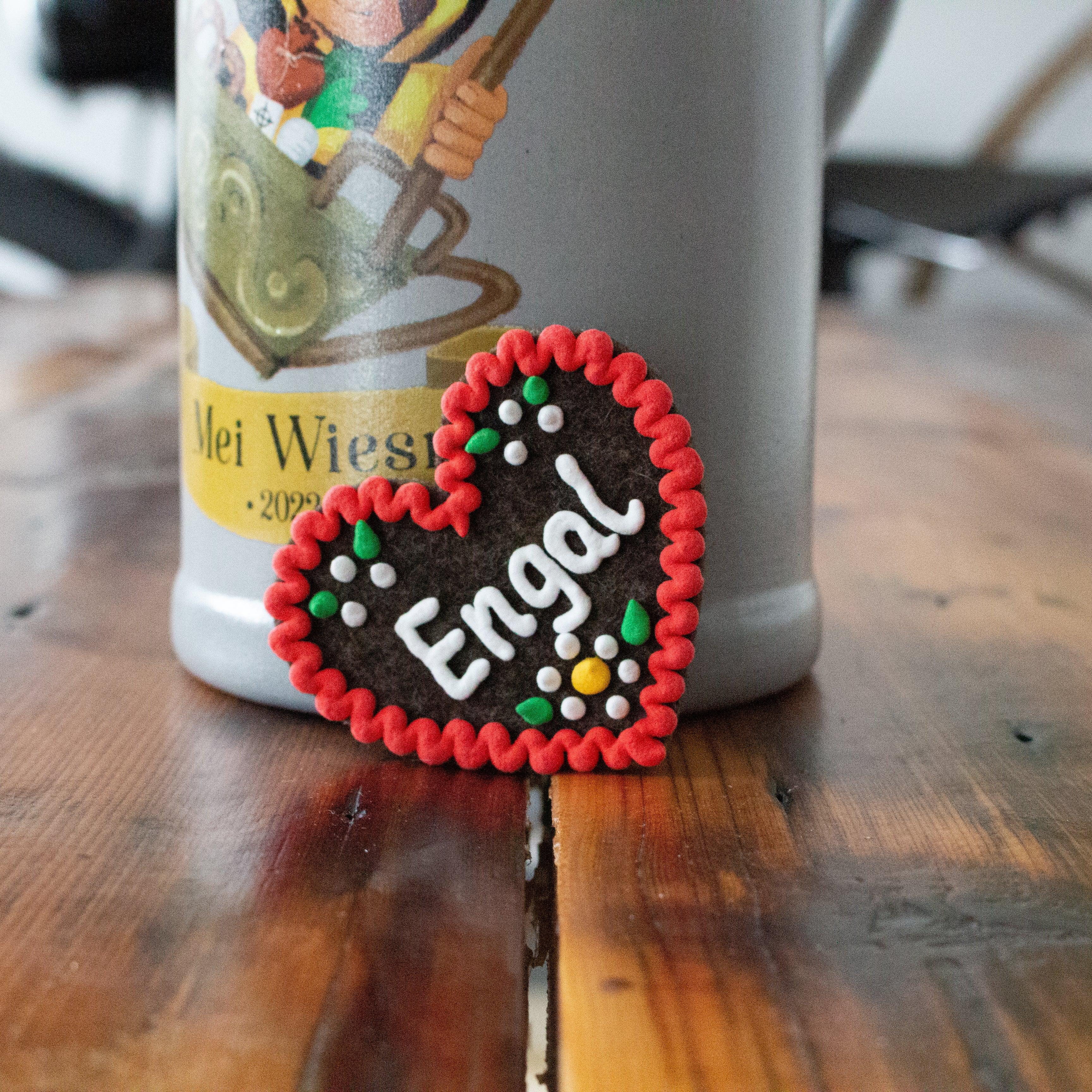 Brauner Filzherz-Anstecker "Engal" mit rotem Rand und weißen, grünen, gelben Dekor-Details vor Bierkrug.