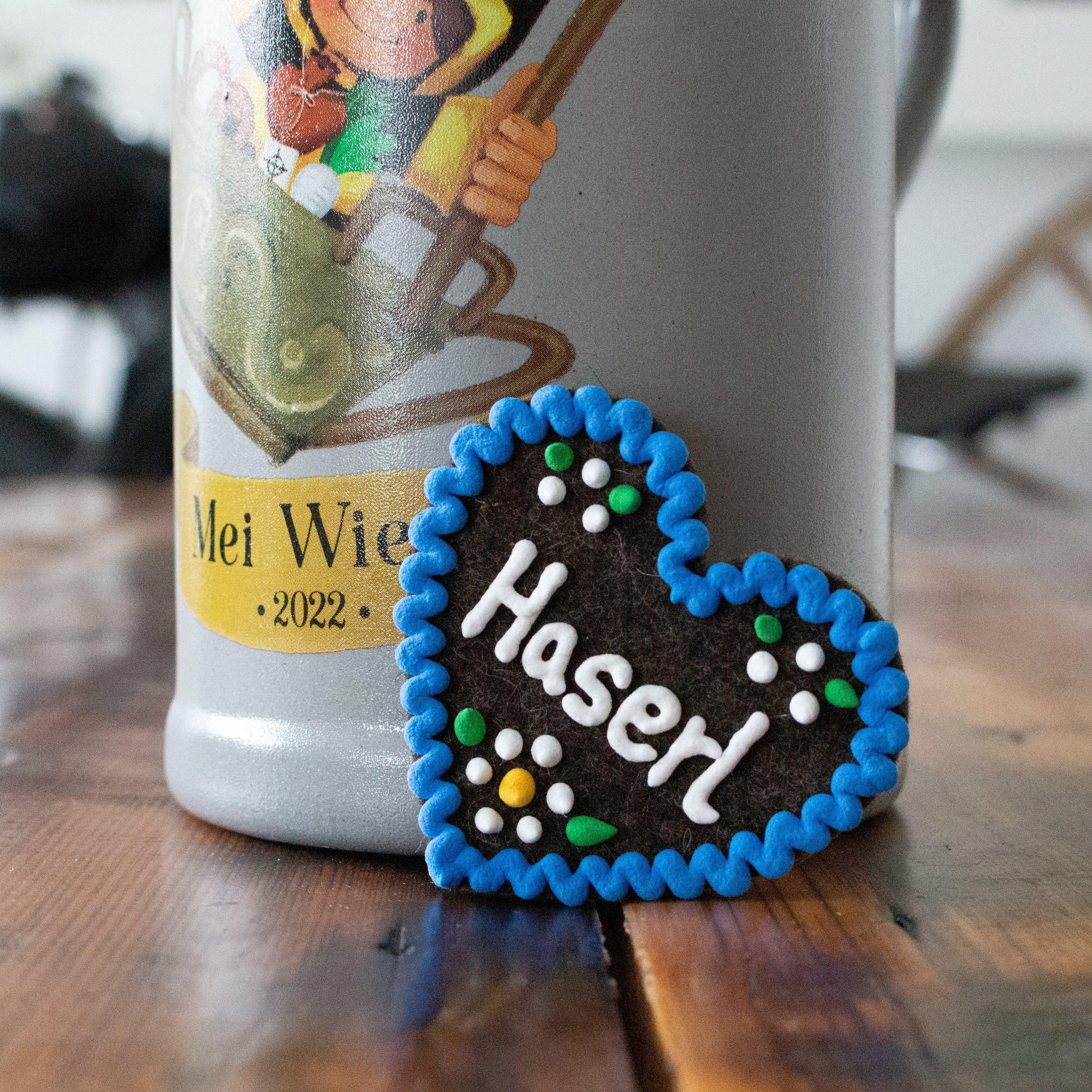 Brauner Filzherz-Anstecker "Haserl" mit blauem Rand und weißer Schrift, dekorativ vor einem Bierkrug auf Holz.