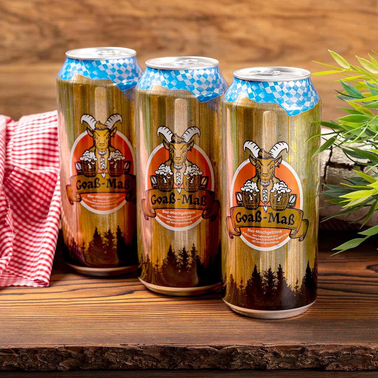 Drei Goaß-Maß XXL-Dosen 1l im bayerischen Stil auf rustikalem Holztisch mit Holz und grünem Tuch.