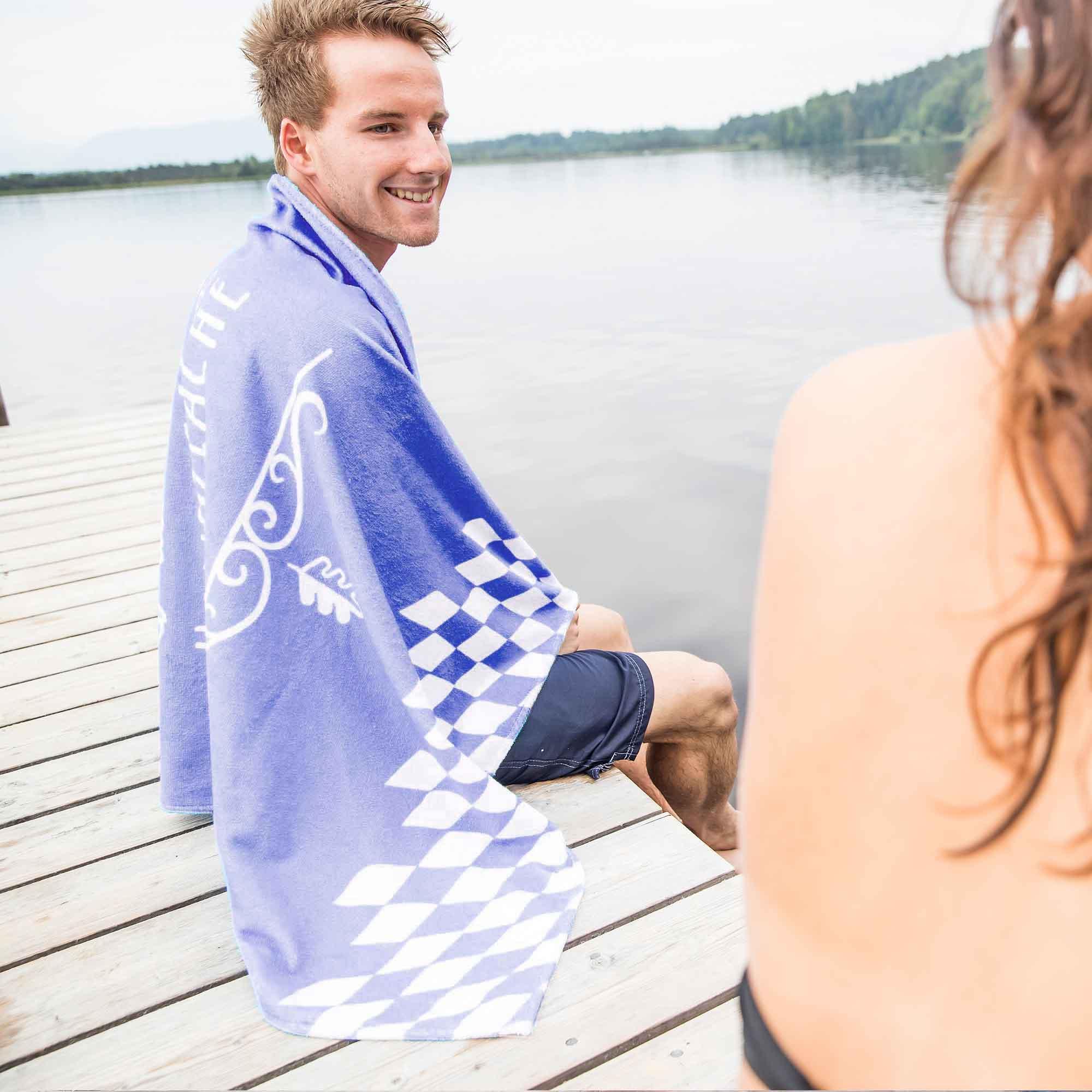 Junger Mann mit blauem bayrischem Badetuch auf Holzsteg am See im Sommerurlaub sitzend