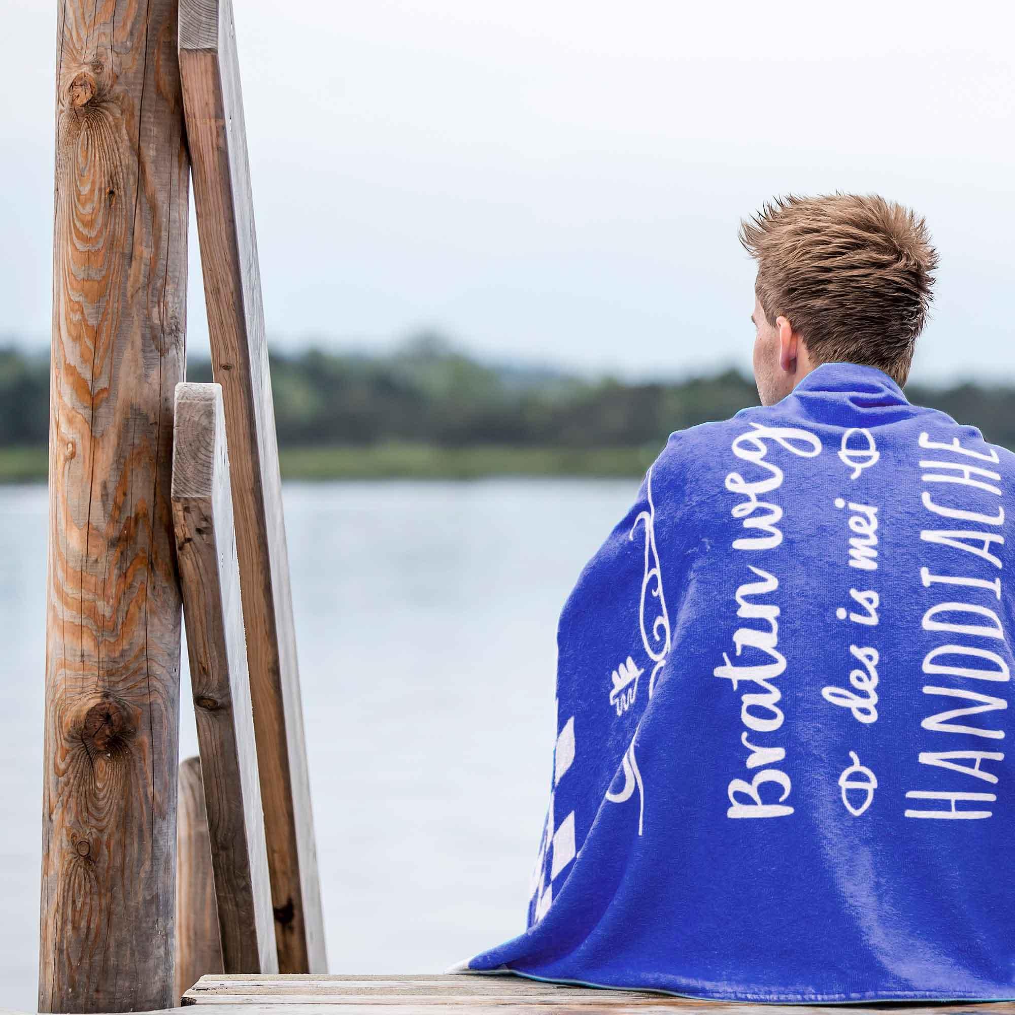 Person von hinten auf Holzsteg am See mit blauem Handtuch mit Aufschrift Bratzn weg des is mei Handdiachl