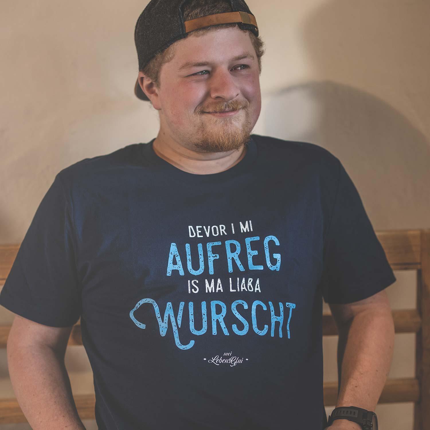 Lachender Mann im Herren T-Shirt "Bevor i mi aufreg...", dunkles Shirt mit blauem Spruch und Baseballcap.