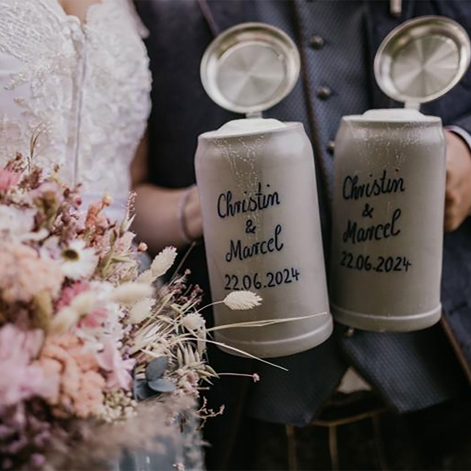 Paar hält zwei gravierte PREMIUM Bierkrug Steinkrüge mit Namen und Hochzeitsdatum, neben Blumen.
