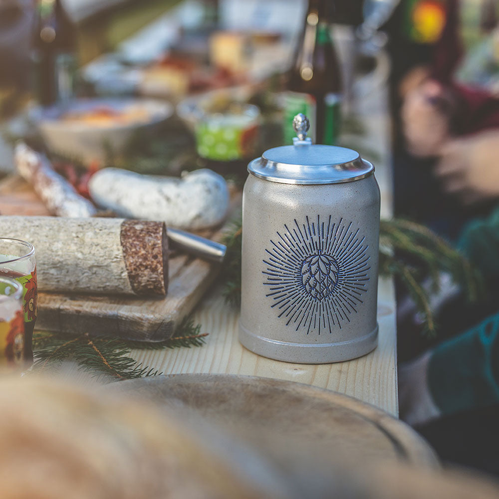 Detailansicht eines Bierkrugs aus Steingut mit blauem Hopfen-Motiv und Zinndeckel auf rustikalem Holztisch.