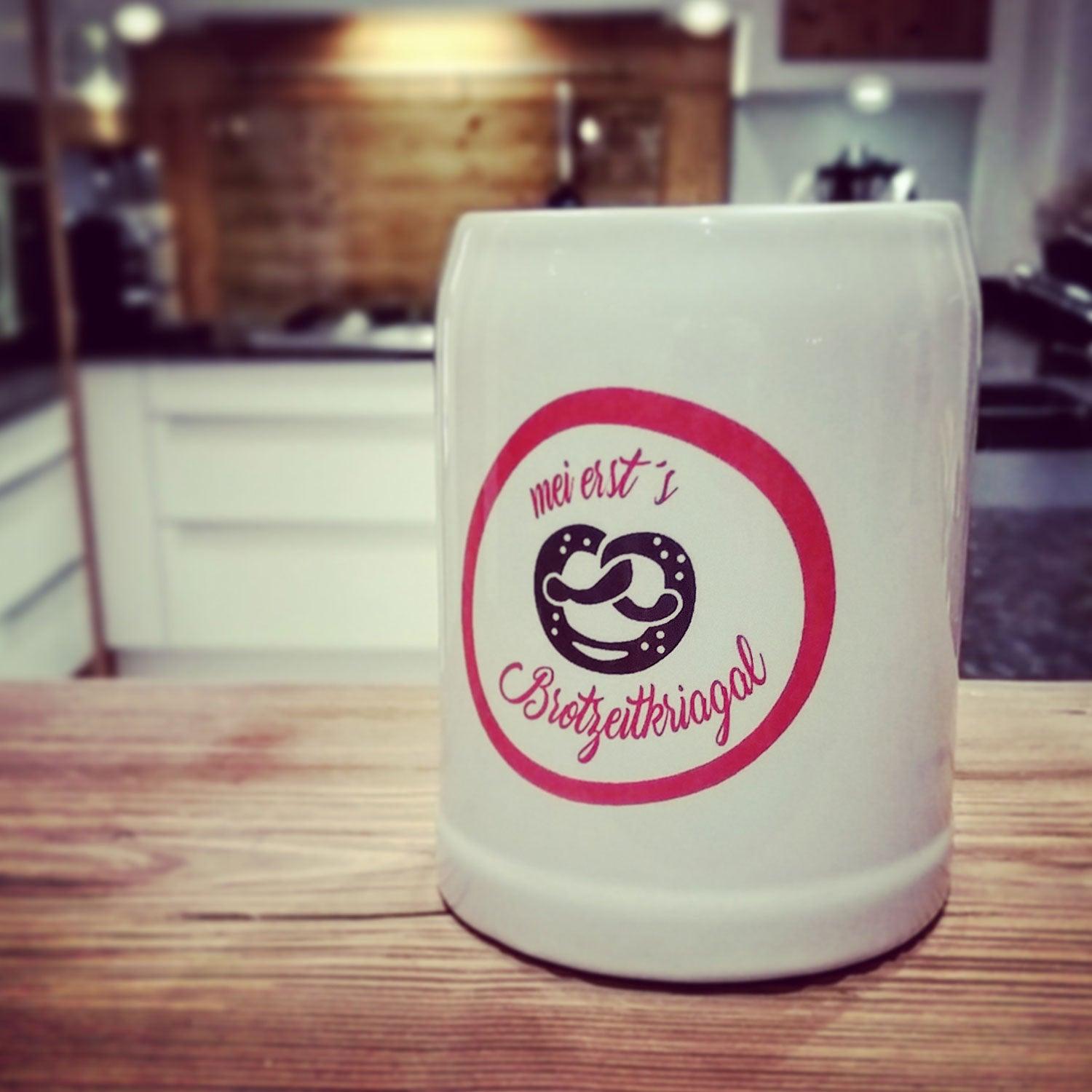 Weißer Brotzeitkrug mit Brezel-Logo und bayerischem Spruch auf rustikalem Holztisch im Shop.