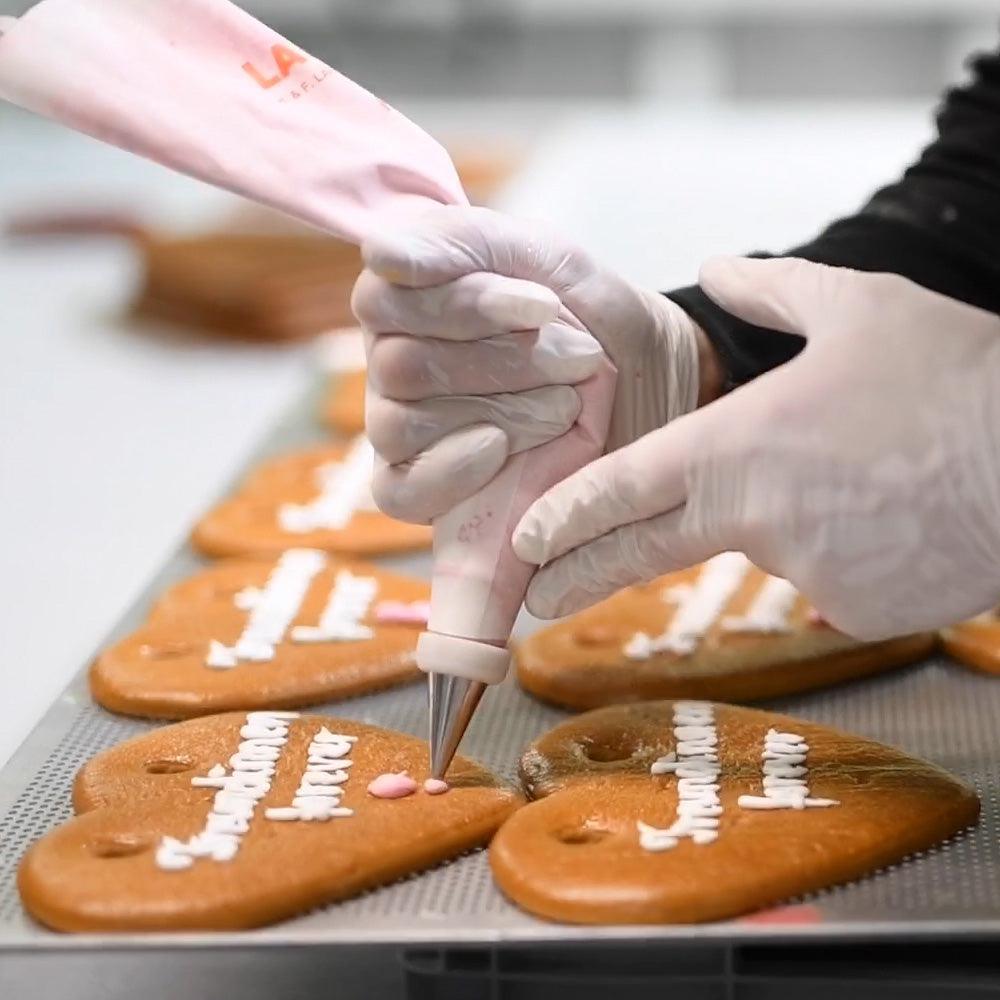 Hände in weißen Handschuhen verzieren Lebkuchenherzen auf Backblech mit rosa und weißer Zuckerguss-Creme.