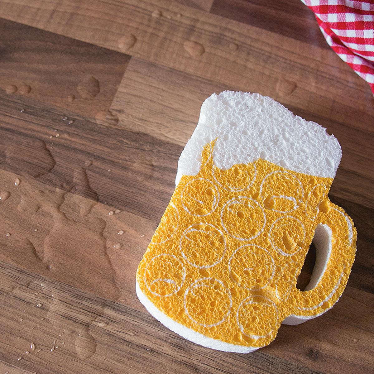 Bierkrugförmiger Küchenschwamm in Gelb und Weiß mit Schaum-Illustration auf Holztisch und kariertem Tuch.