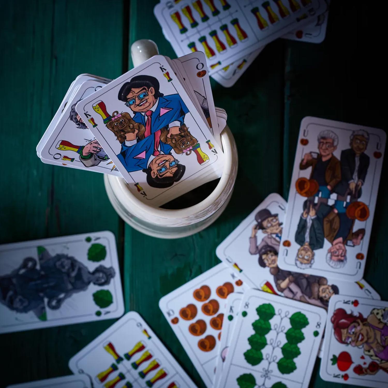 Mehrere Münchner Schafkopf Karten mit bayerischen Motiven und charakteristischen Figuren auf grünem Holztisch ausgebreitet.