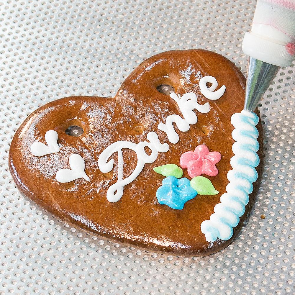 Verzieren eines Lebkuchenherzens mit weißer Zuckerglasur und bunten Blumen, Schriftzug "Danke", Backblech-Hintergrund.