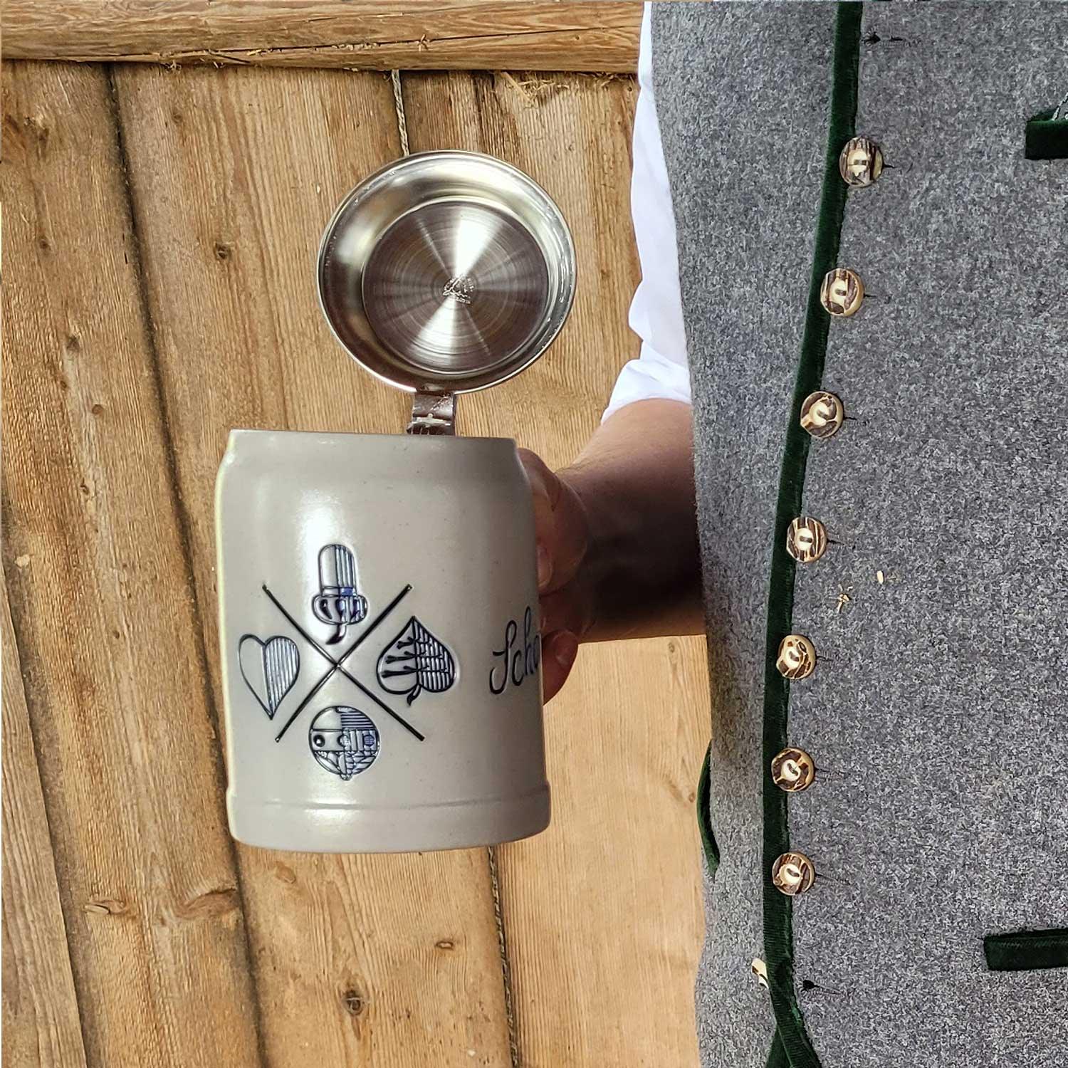 PREMIUM Bierkrug Bayrisch Blatt mit Zinndeckel, traditionellen Symbolen und "Schöner" Schriftzug in Hand eines Mannes mit Trachtenweste.