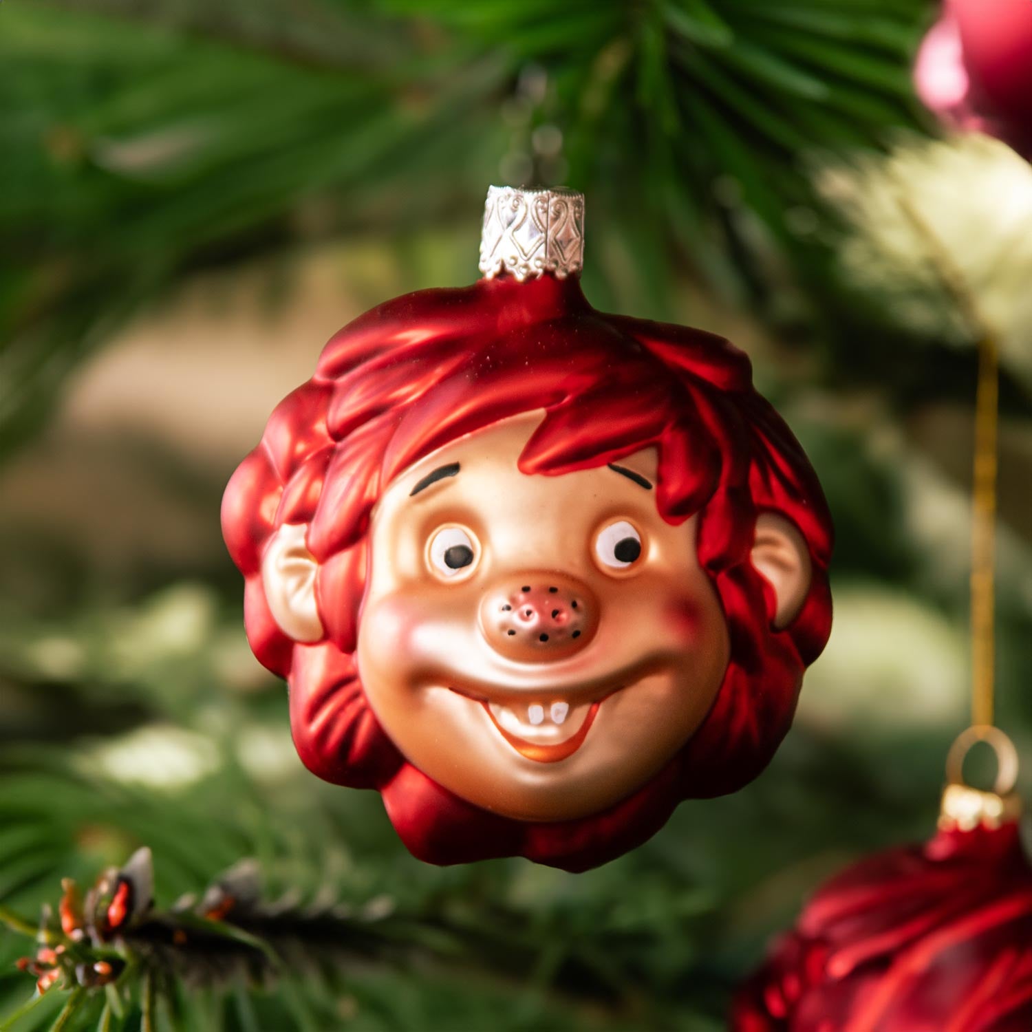 Pumuckl Weihnachtskugel aus Glas, rot-goldene Figur des Kobolds mit breitem Lächeln im Weihnachtsbaum.