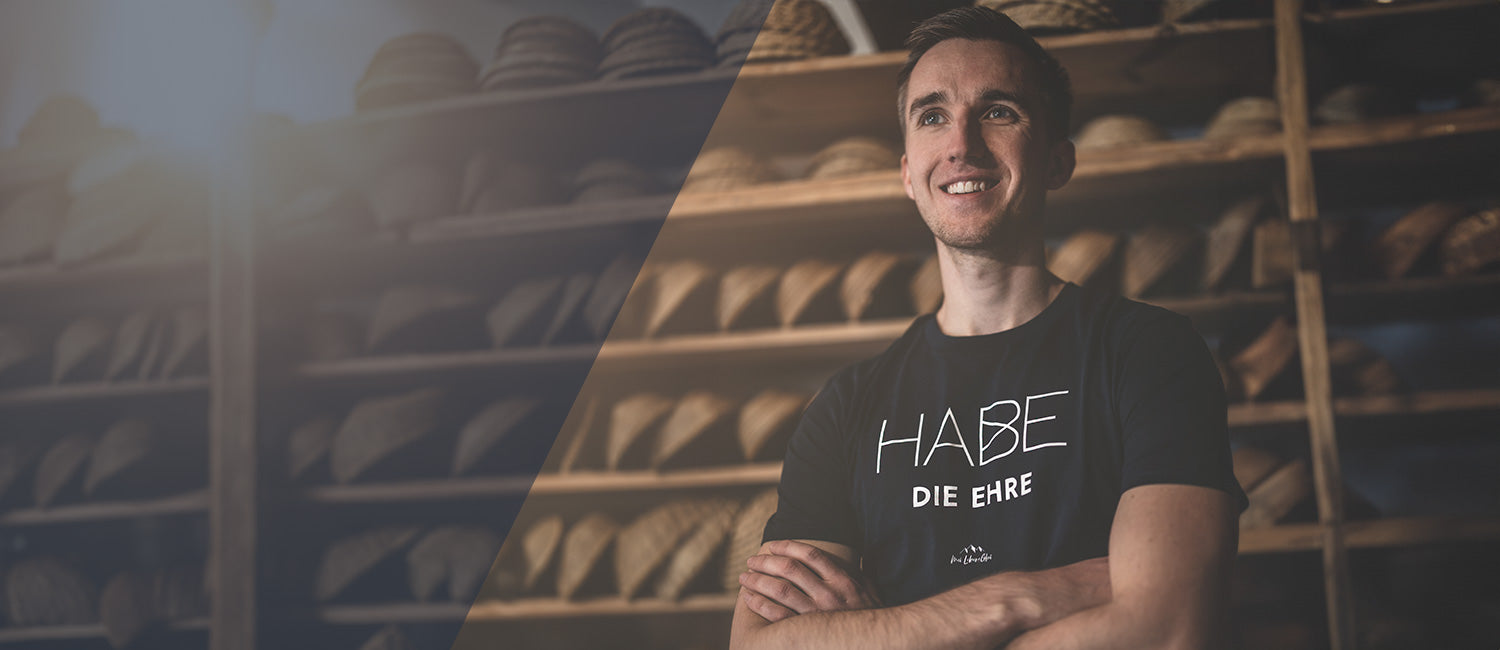 Junger Mann mit dunklem Habe Die Ehre T-Shirt, freundlich lächelnd, vor rustikalen Holzregalen. Authentisches bayerisches Herren Shirtdesign.