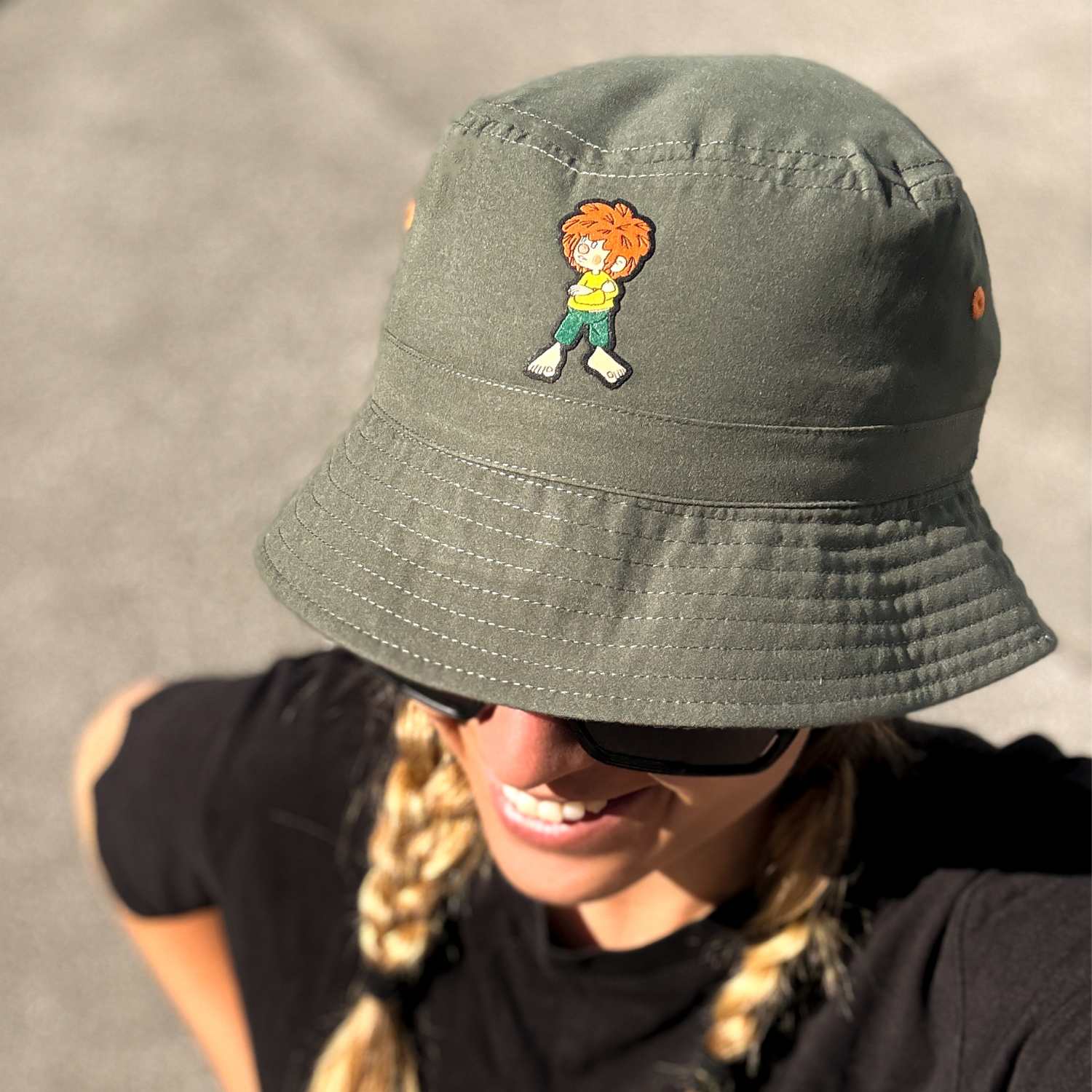 Pumuckl Fischerhut "Grantla" in Khaki mit Pumuckl-Emblem auf dem Kopf einer lächelnden Frau.