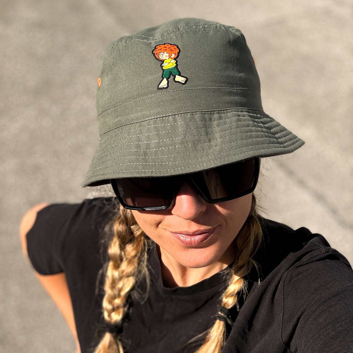 Frau mit blonden Zöpfen trägt einen grünen Pumuckl Fischerhut "Grantla" und Sonnenbrille, von oben fotografiert.