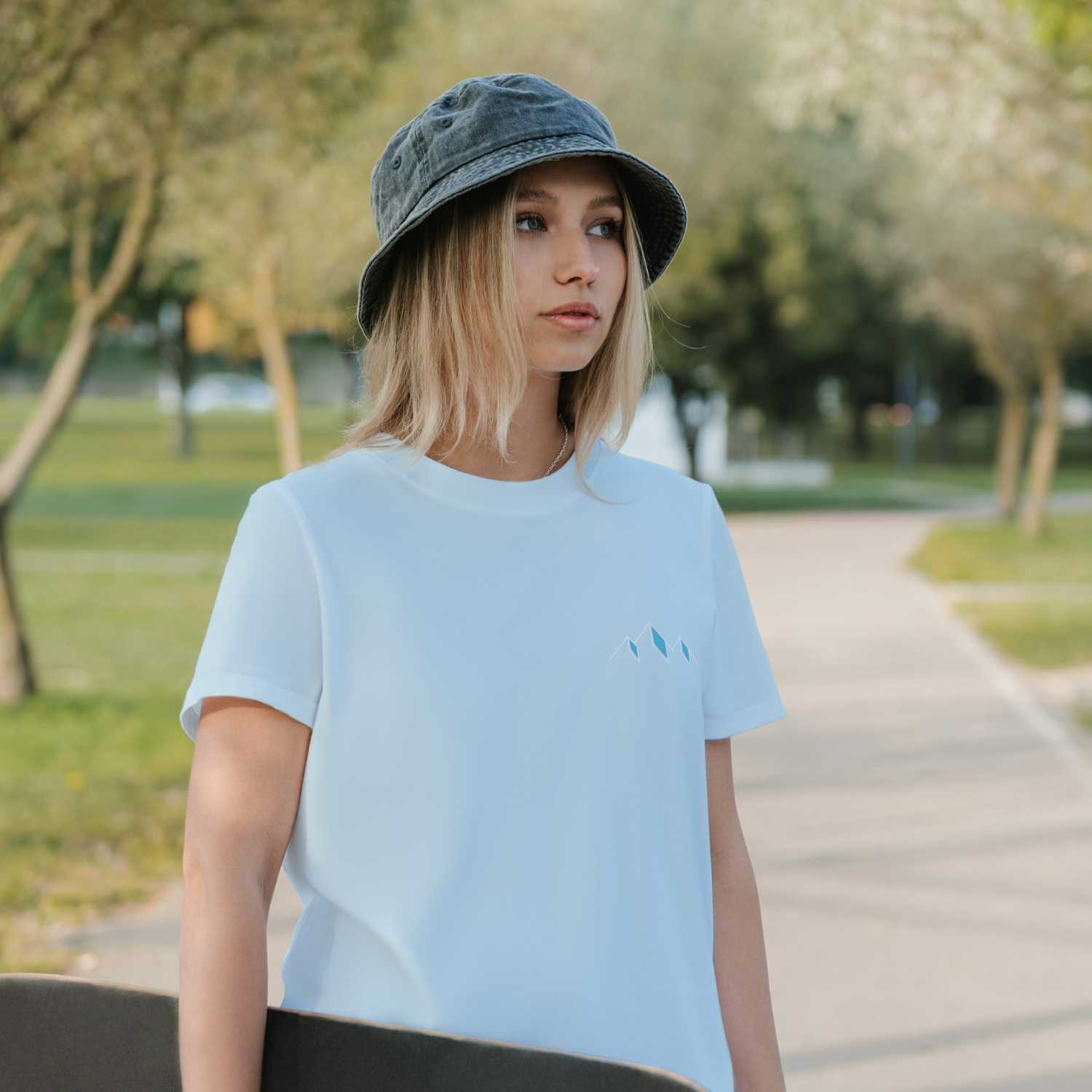 Junge Frau trägt weißes Damen T-Shirt "Himmi Herrgott" mit blauem Bergmotiv in Parkumgebung, Eimerhut, Skateboard.