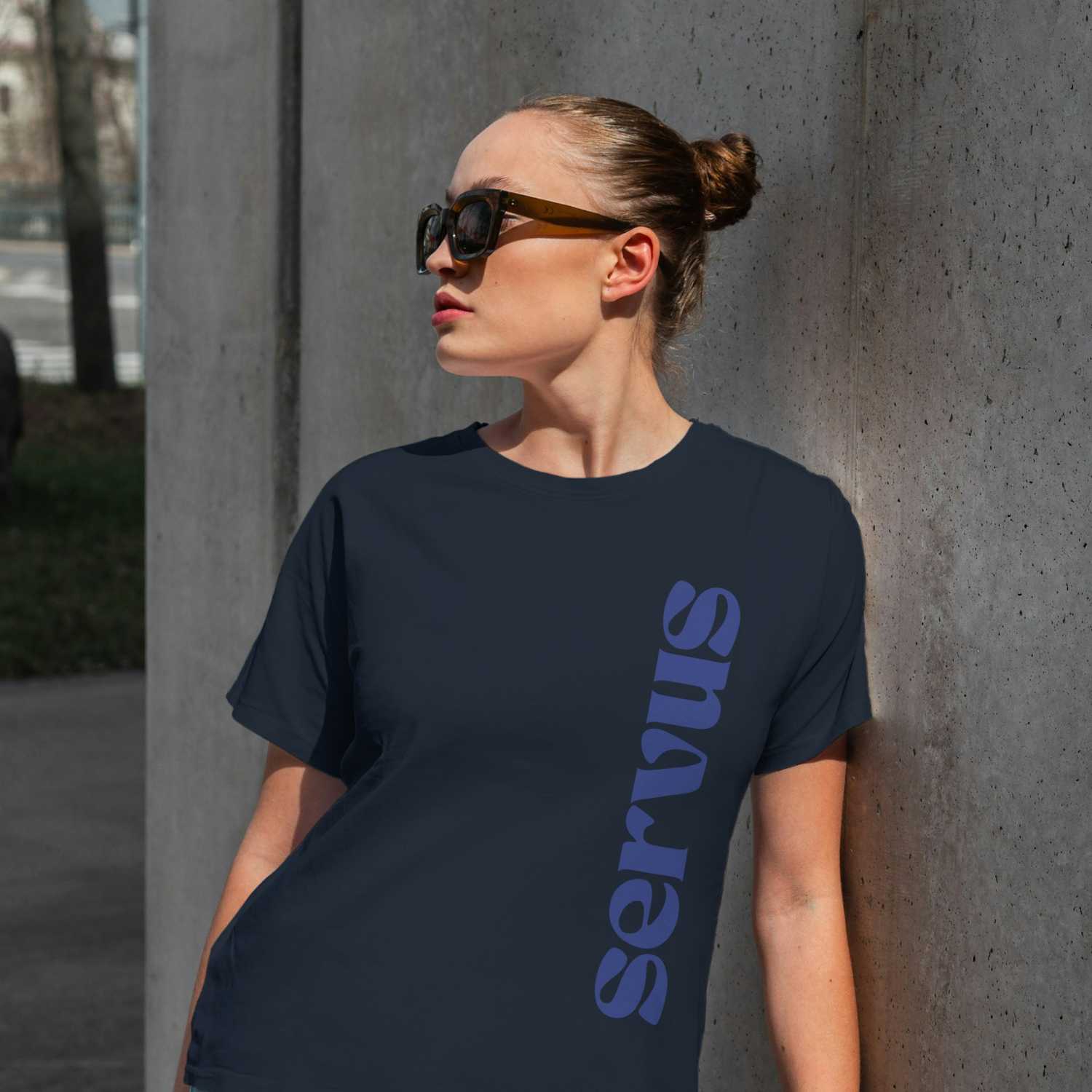Model trägt ein dunkelblaues Damen T-Shirt Servus mit lila Schrift, lässiger Freizeitschnitt, schwarze Sonnenbrille.