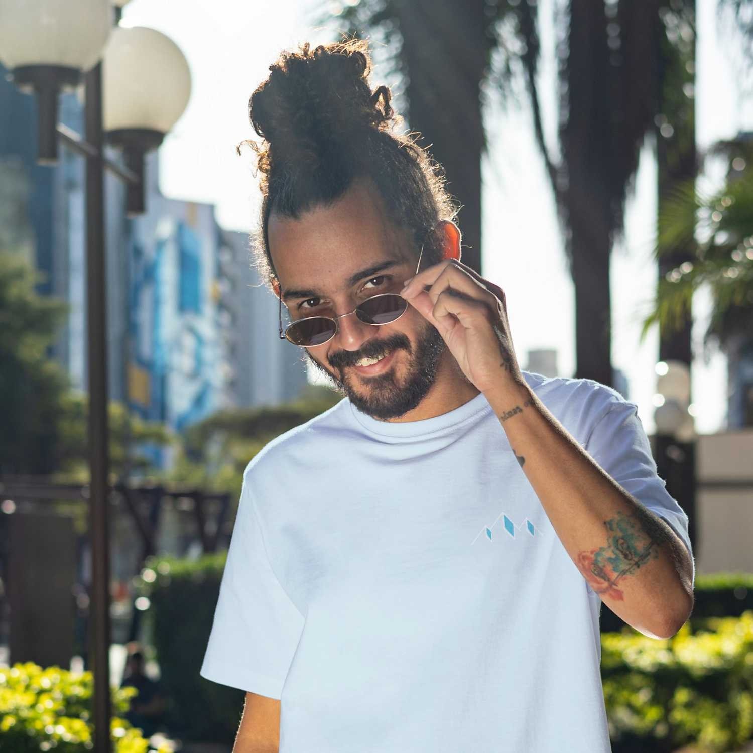 Junger Mann mit Dutt und Bart, trägt blaues Herren T-Shirt "Himmi Herrgott" mit stilisiertem Berglogo, Sonnenbrille leicht gesenkt.