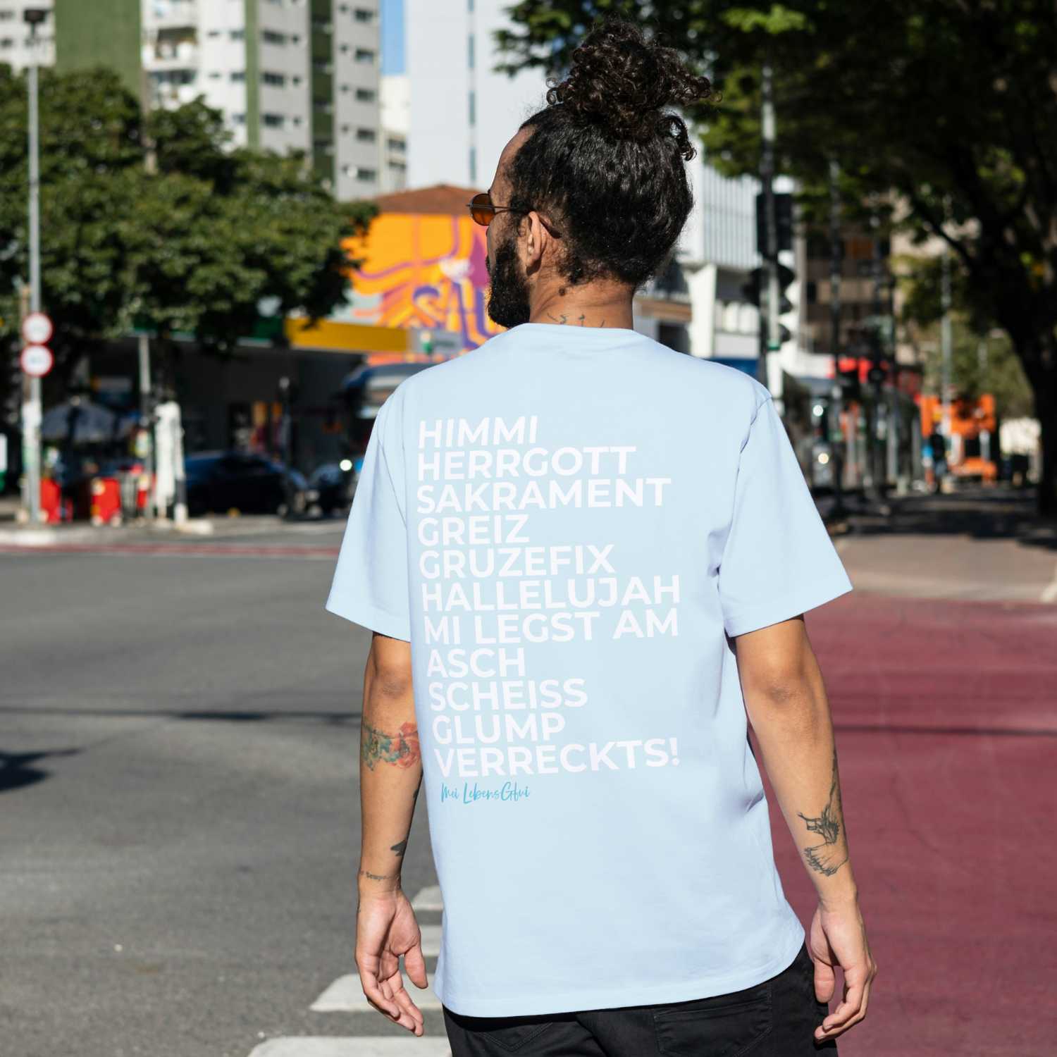 Rückenansicht eines Mannes in hellem Herren T-Shirt "Himmi Herrgott" mit bayerischen Ausrufen auf der Straße stehend.
