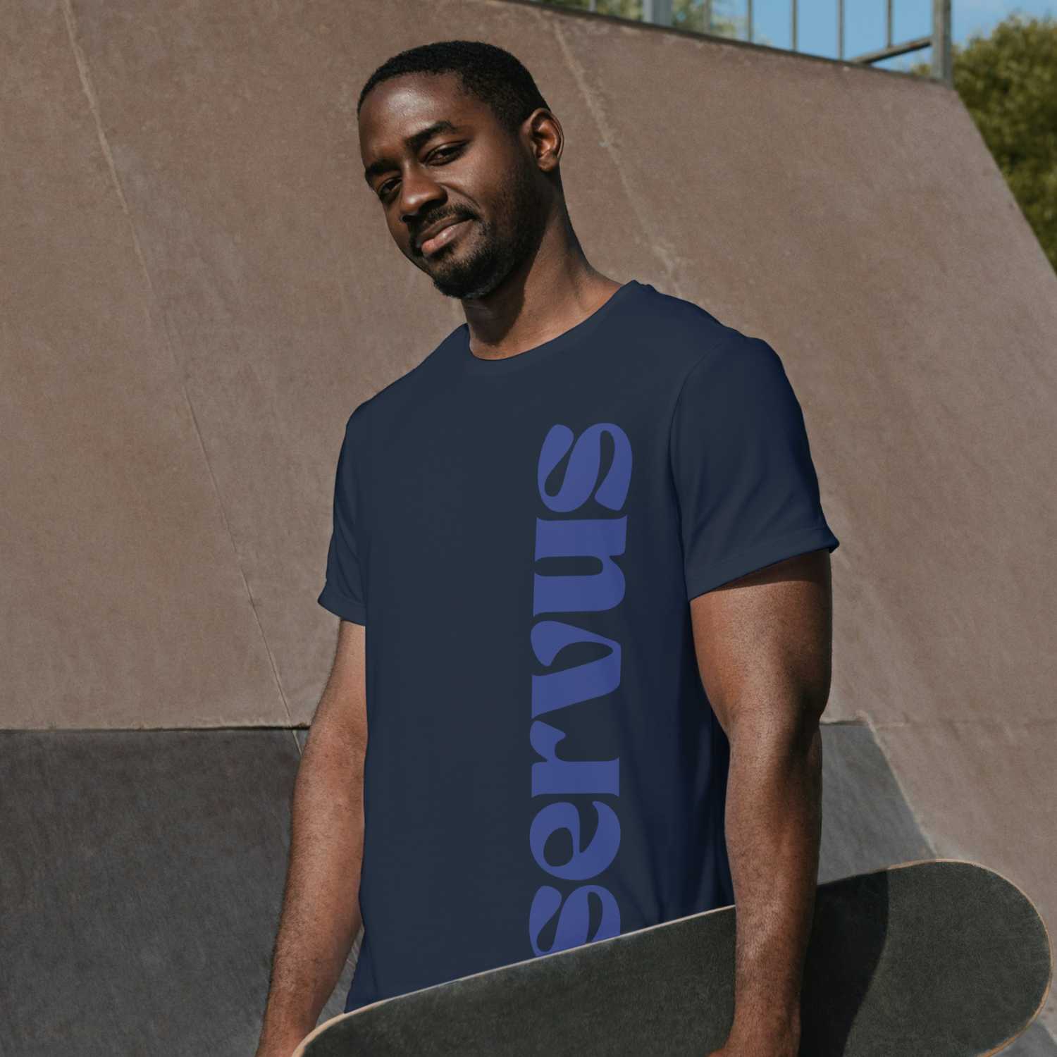 Lächelnder Mann im dunkelblauen Herren T-Shirt Servus mit Skateboard auf einem Skatepark.