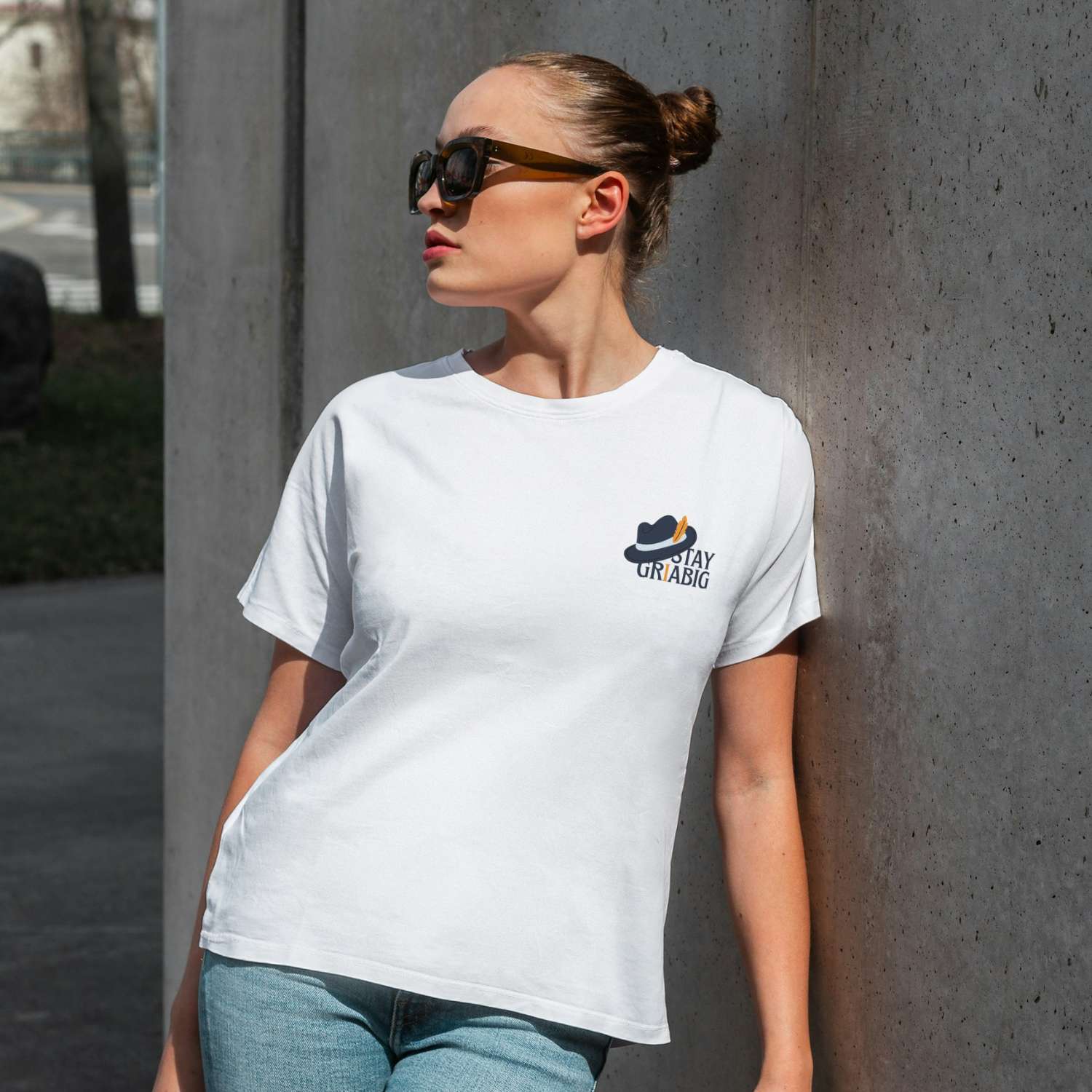 Model trägt weißes Damen T-Shirt Stay Griabig mit kleinem Hut-Logo, dunkler Sonnenbrille, und Jeans im Freien.