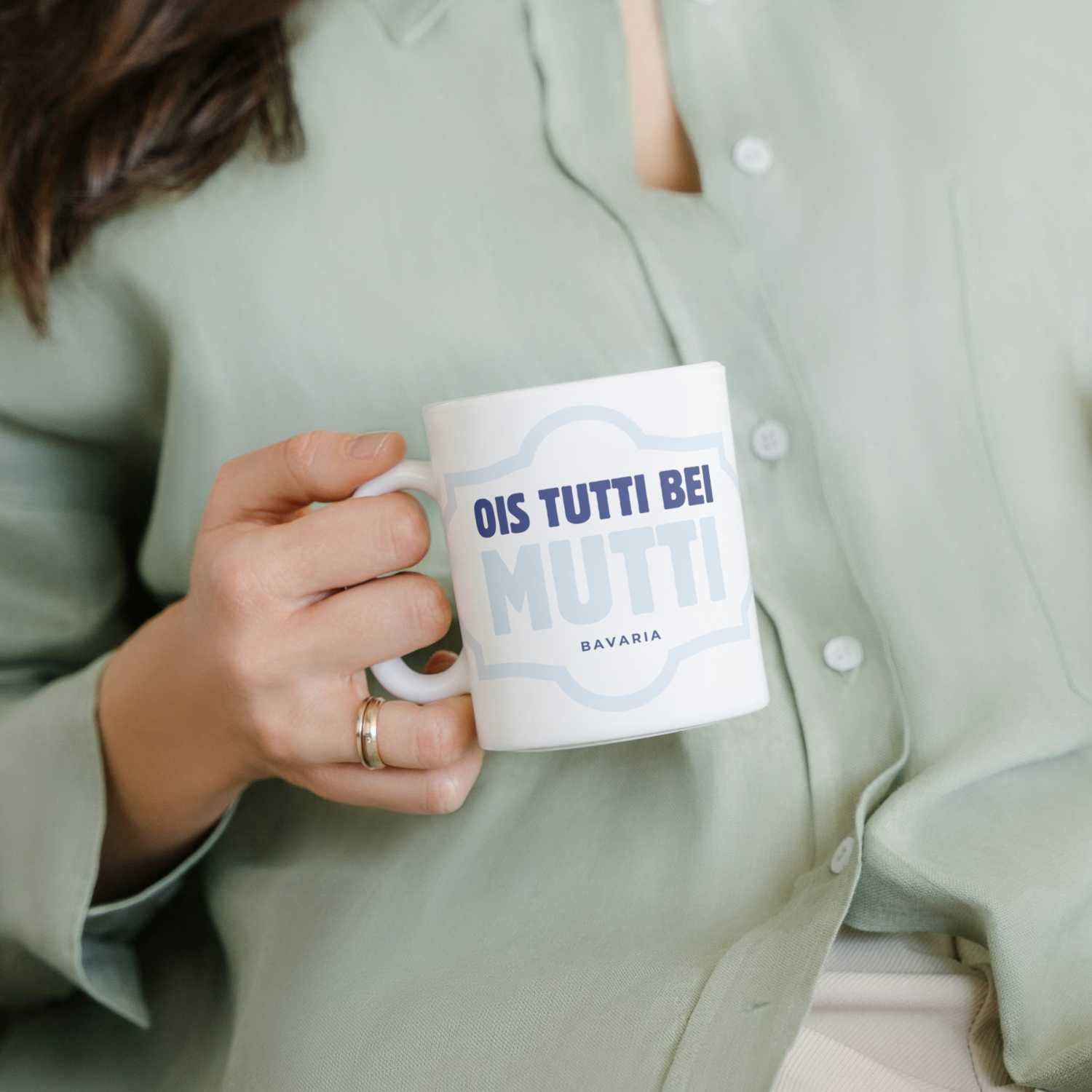 Hand hält weißes Kaffee Haferl Ois tutti bei Mutti mit blauem Text, Frau trägt grüne Leinenbluse.