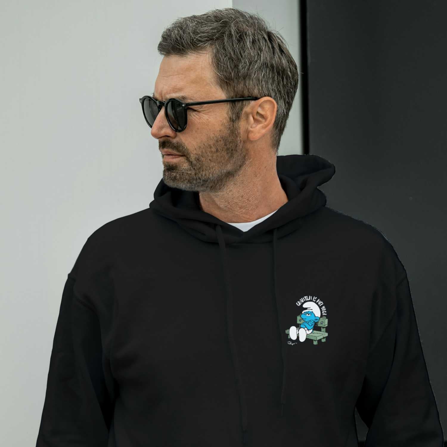Mann mit Sonnenbrille trägt schwarzen Schlümpfe Herren Hoodie "Grantln is mei Yoga" mit Grantel-Schlumpf Motiv.