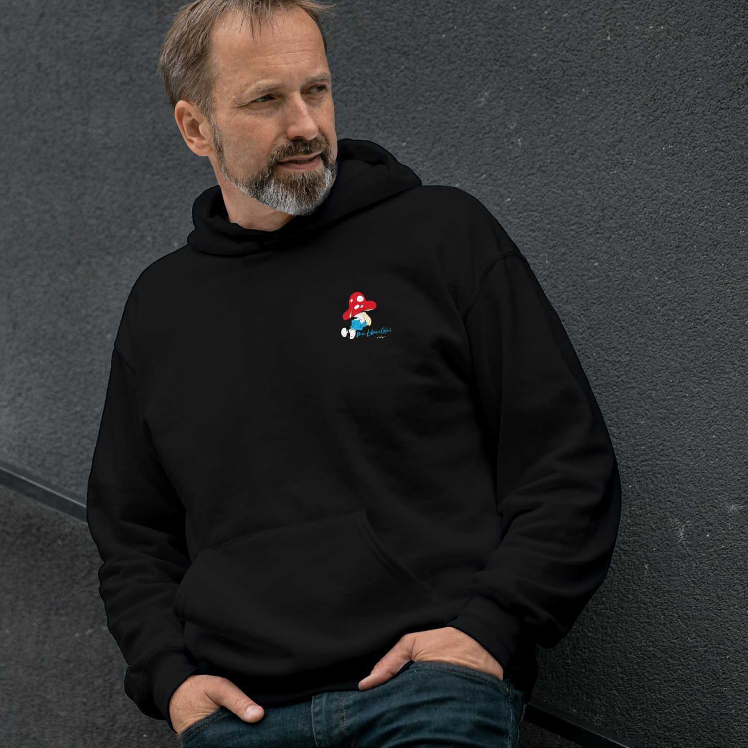 Ein Mann trägt einen schwarzen ®Die Schlümpfe Herren Hoodie Do legst di nieda mit Pilz-Schlumpf-Print.