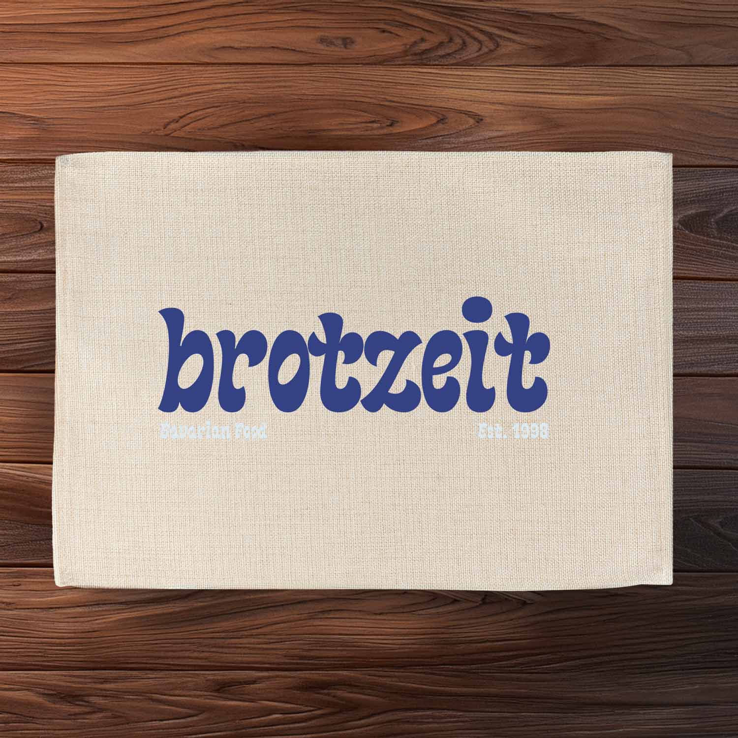 Beiges Tischset Brotzeit est. 1998" mit blauem Schriftzug und "Bavarian Food" Aufdruck auf Holztisch.
