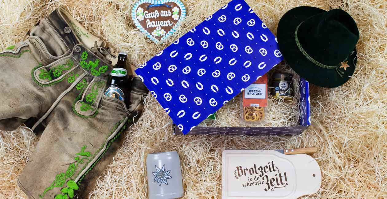 Bayrisches Geschenkset mit Lederhose, Bier, Filzhut, Lebkuchenherz und Brotzeit-Zubehör auf Heu.