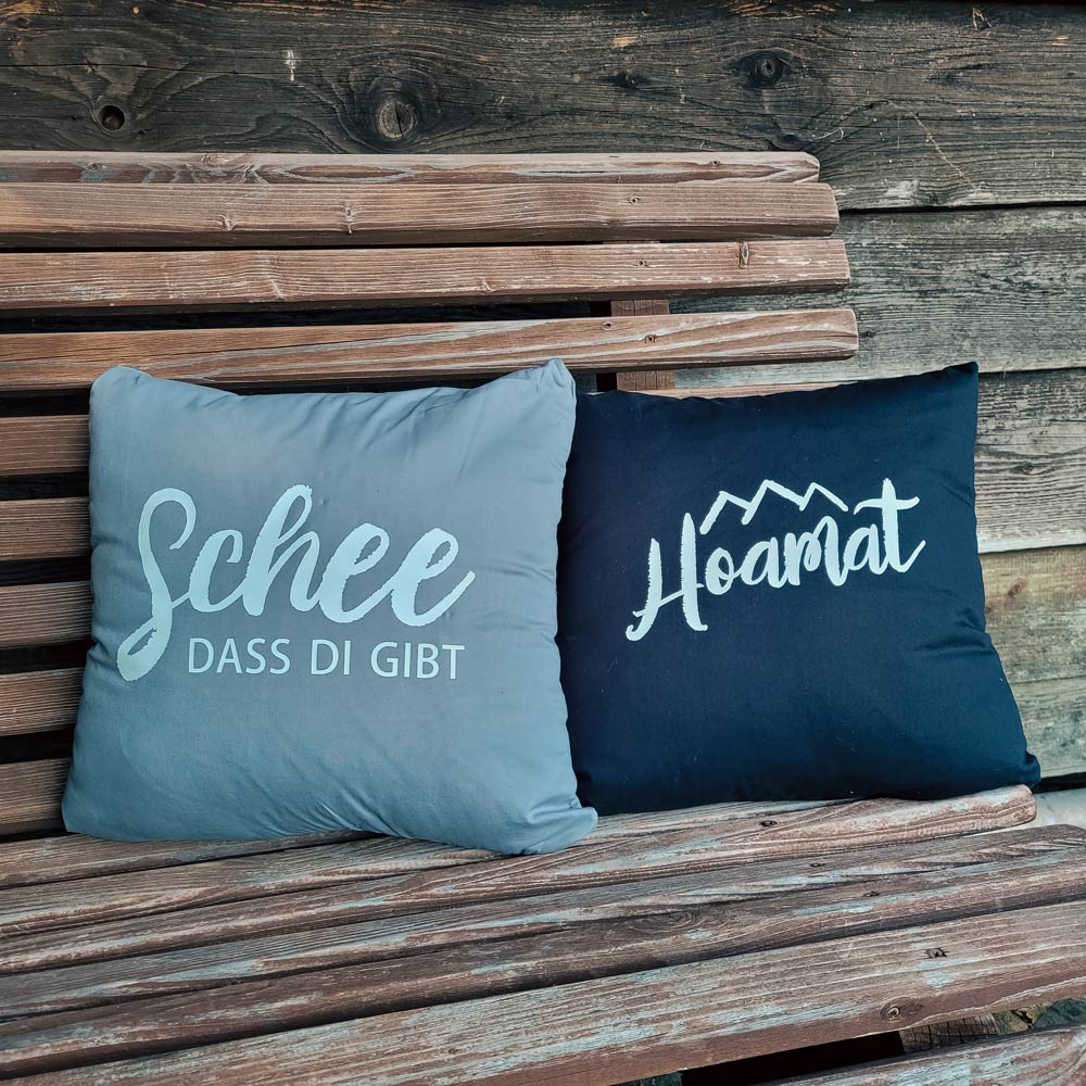 Zwei Zierkissen mit schöner Typografie auf einer rustikalen Holzbank: grau mit "Schee dass di gibt" und dunkelblau mit "Hoamat", Trachtenaccessoires.