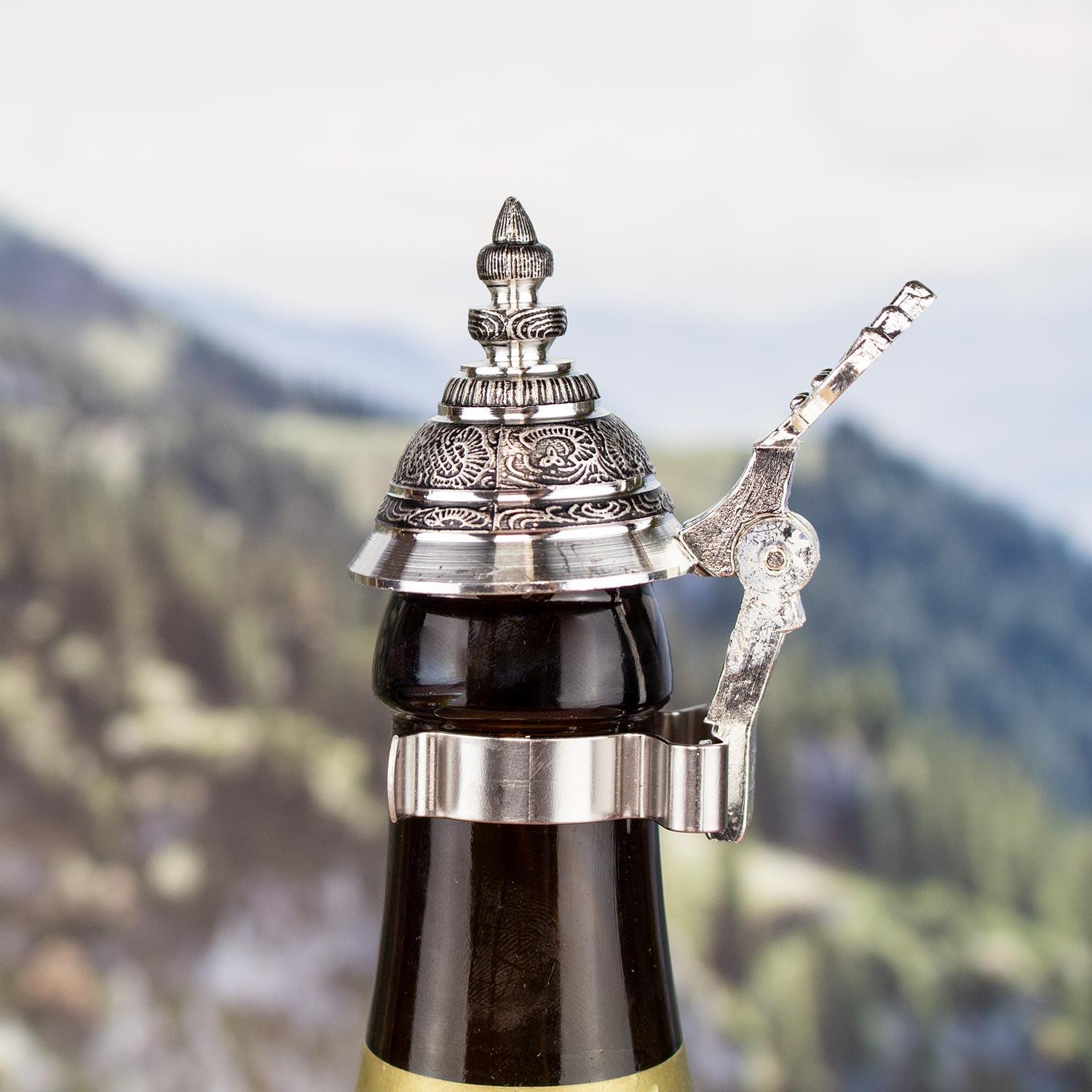 Handgefertigter Zinndeckel mit Verzierungen auf einer braunen Bierflasche vor Alpenkulisse.