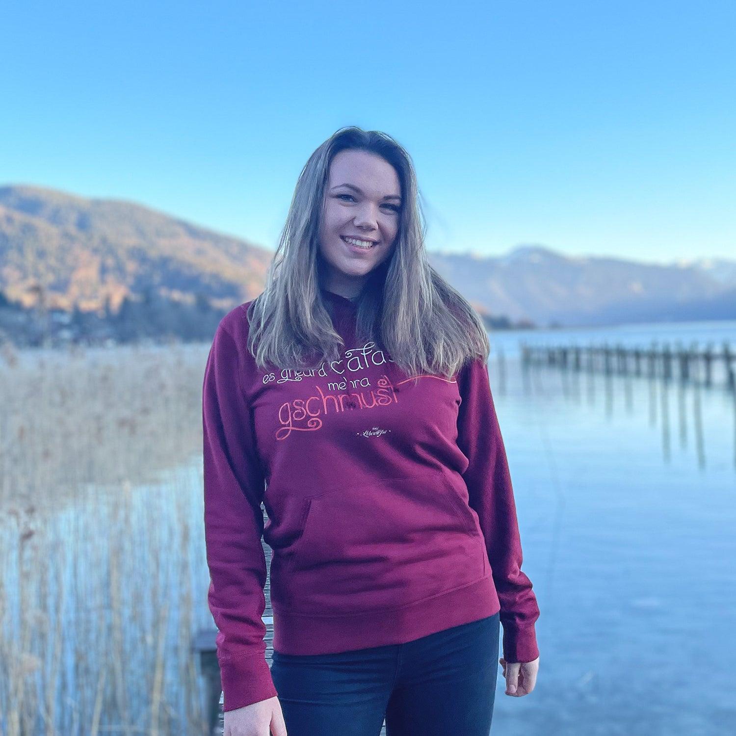 Junge Frau in bordeauxrotem Damen Hoodie Gschmust vor malerischer Seenlandschaft mit Bergen