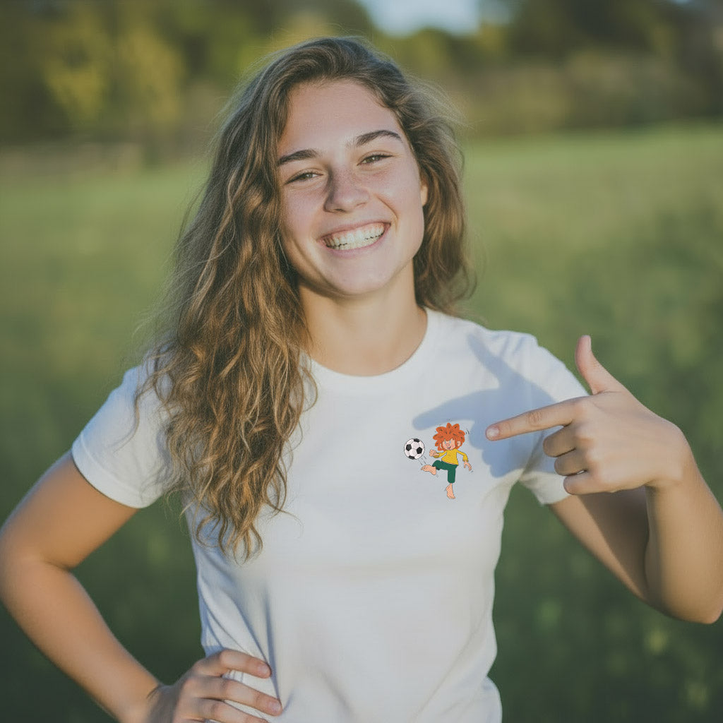 Lächelnde Frau in weißem Pumuckl Damen T-Shirt "Fußball Kobold" auf grüner Wiese zeigend auf Motiv.