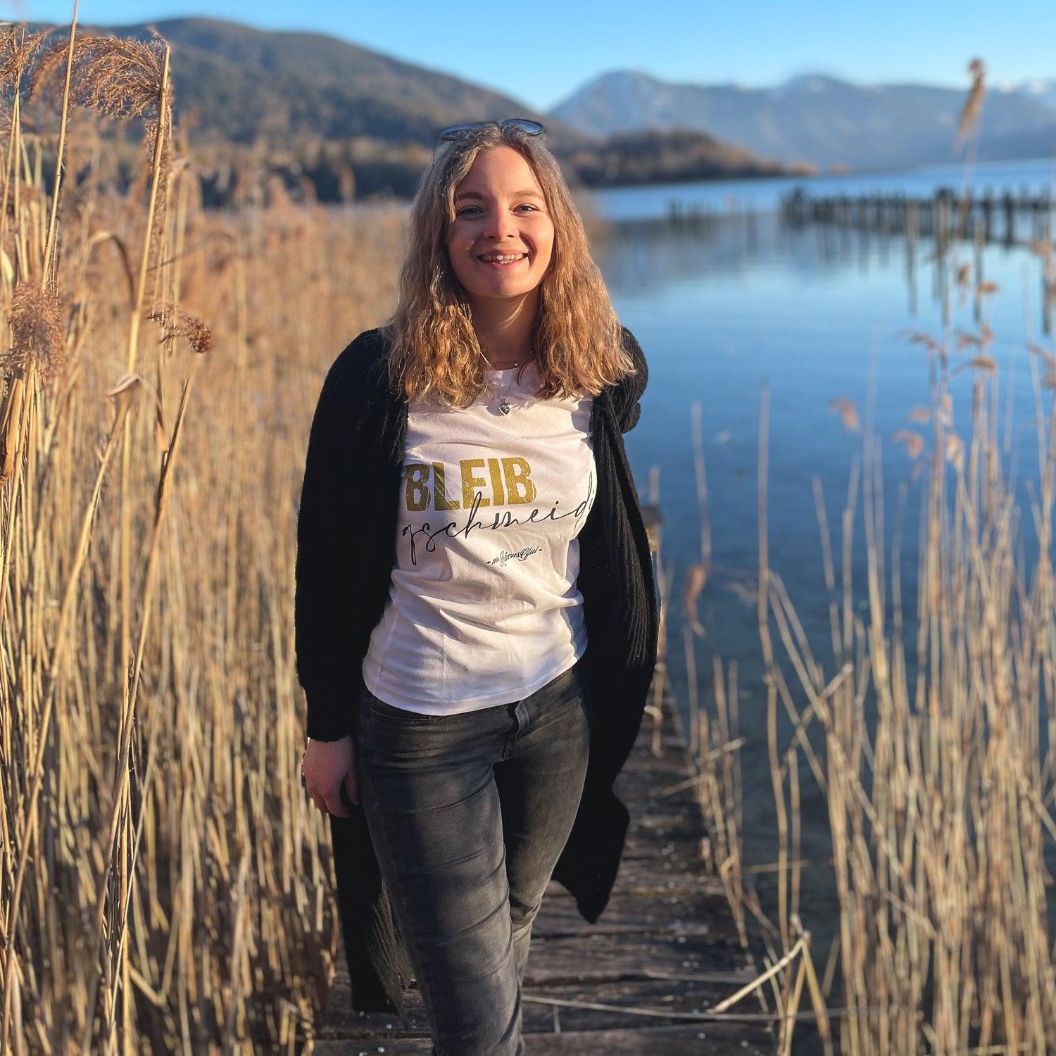 Junge Frau mit langen blonden Haaren trägt weißes Damen T-Shirt "Bleib gschmeidig" auf Holzsteg am See.