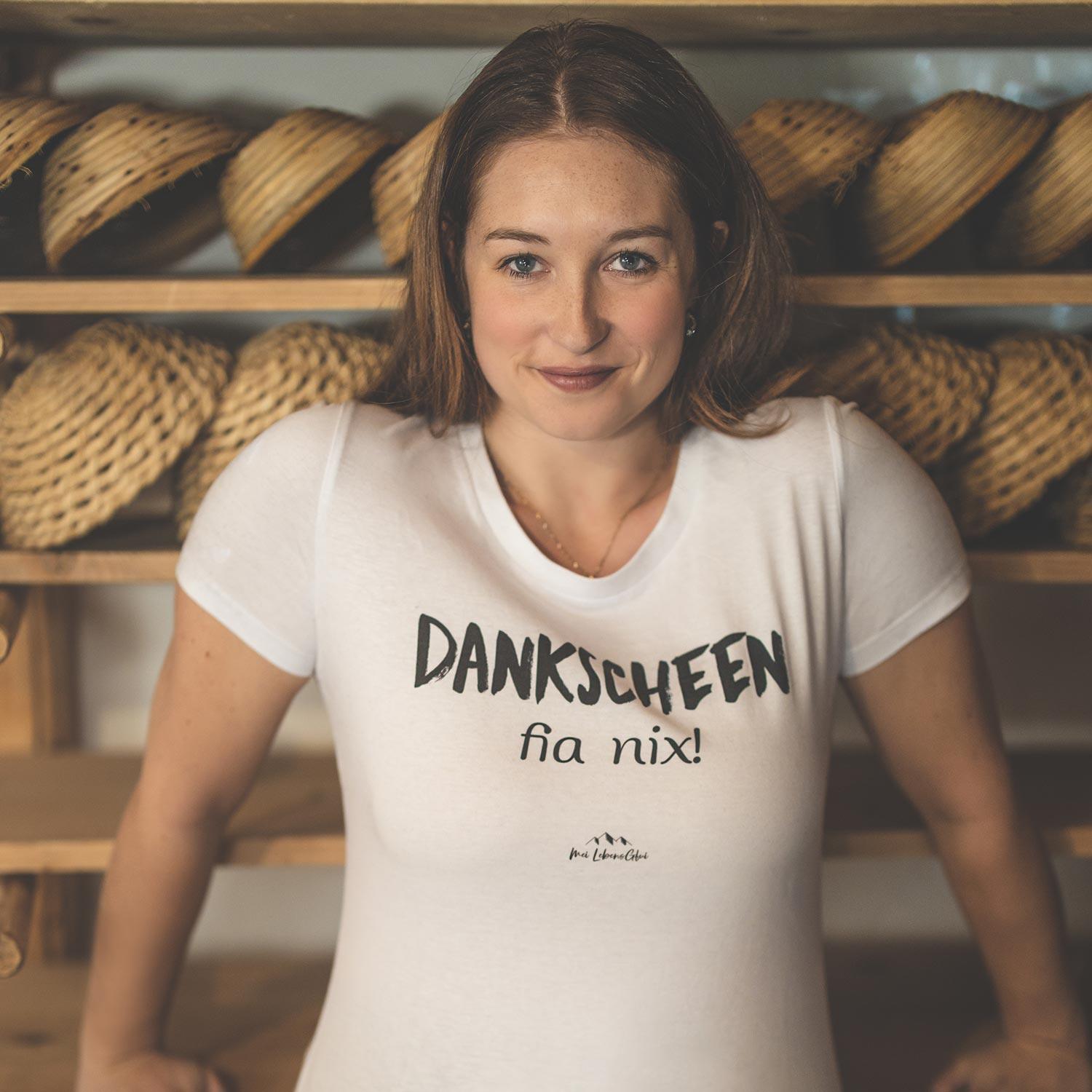 Lachende Frau trägt weißes Damen T-Shirt Dankscheen - fia nix! mit schwarzem Aufdruck vor Holzregal.